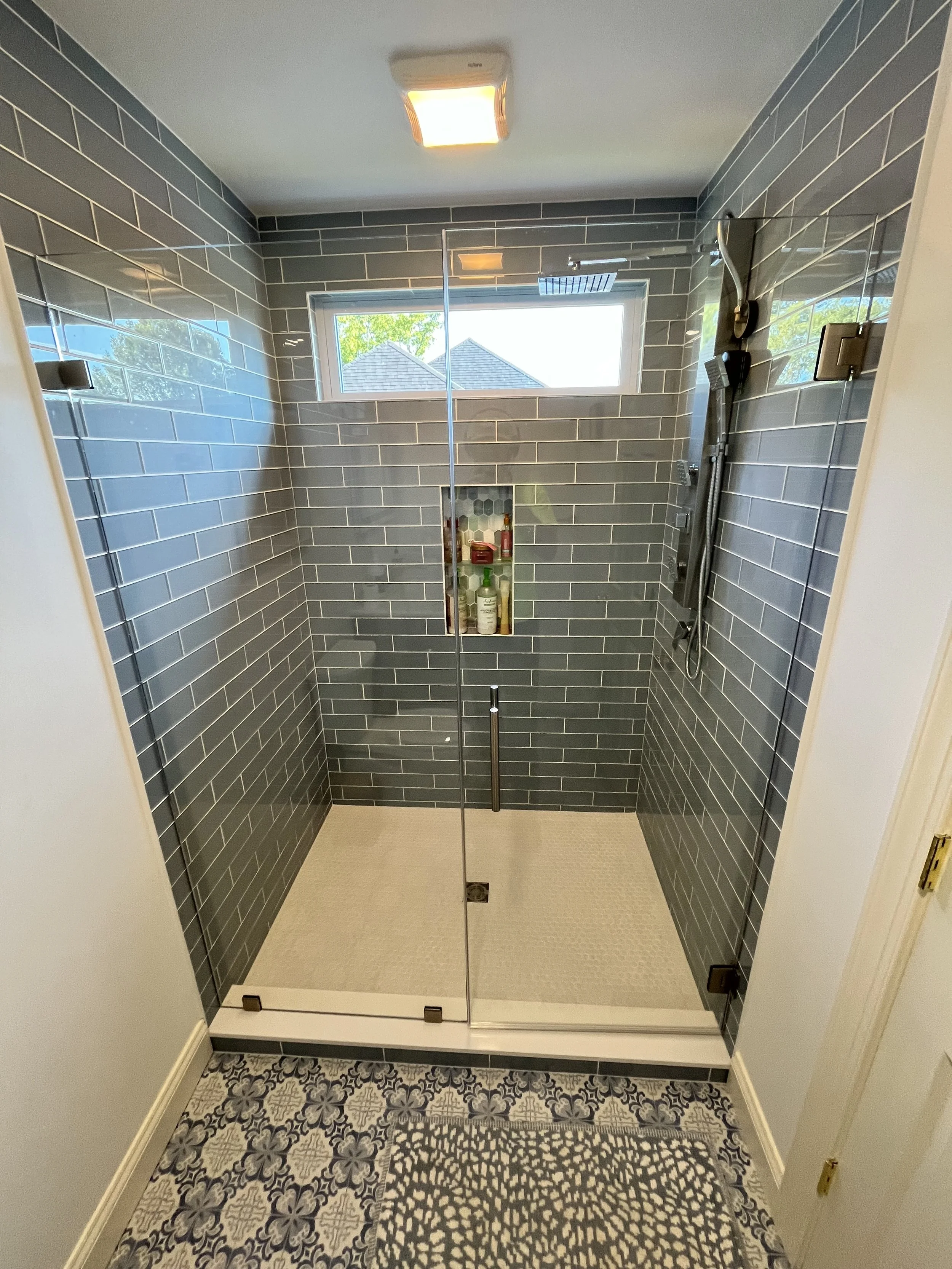 A modern walk-in shower with dark gray subway tiles, a recessed shower niche with bottles of shampoo and body wash, a window showing rooftops and trees outside, a ceiling light, and a patterned tile floor.