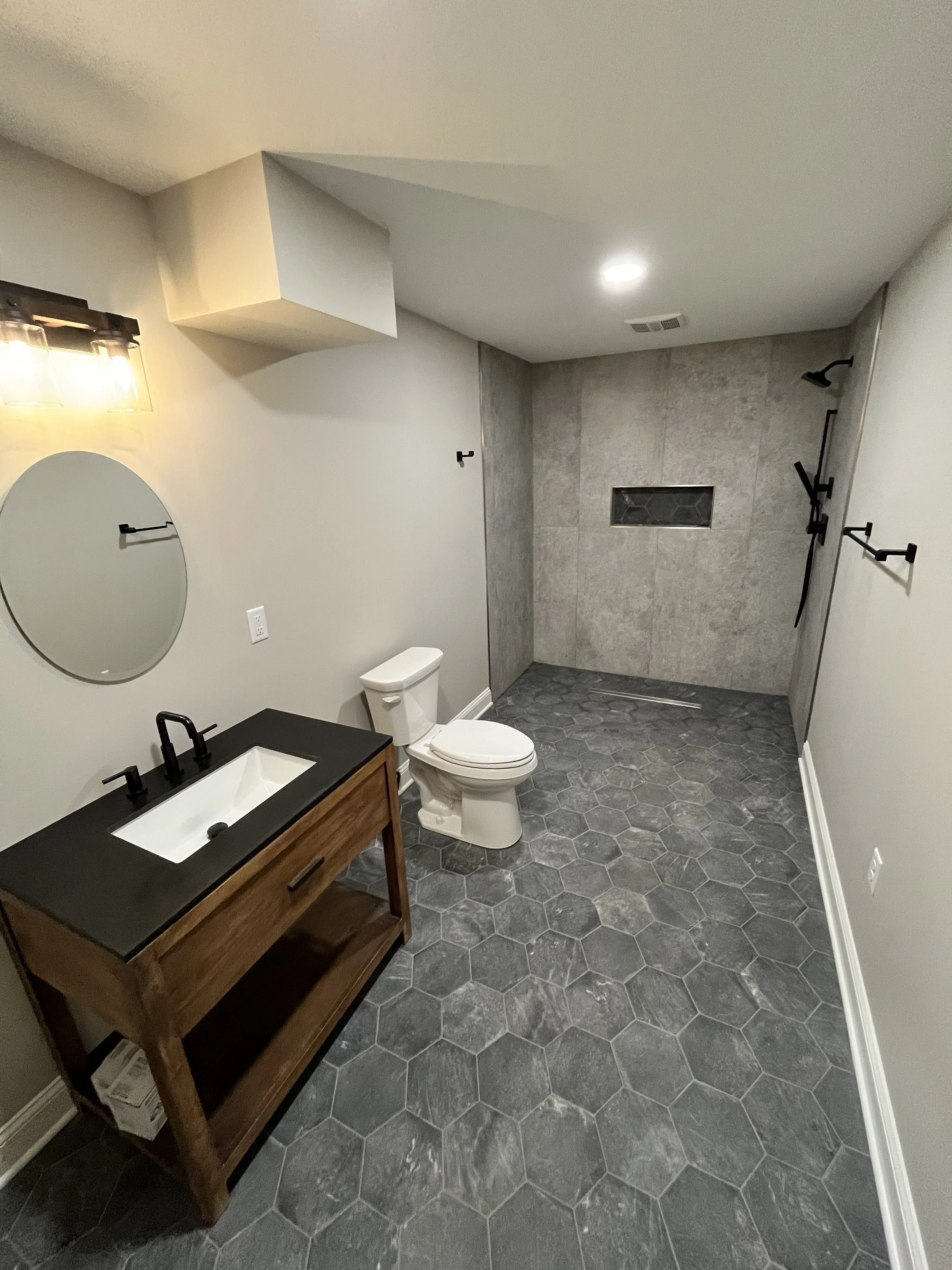 A modern bathroom with dark hexagonal floor tiles, a wooden vanity with a black countertop and white sink, a mirror, a toilet, and a walk-in shower with gray tiled walls and black fixtures, including a showerhead and handles.