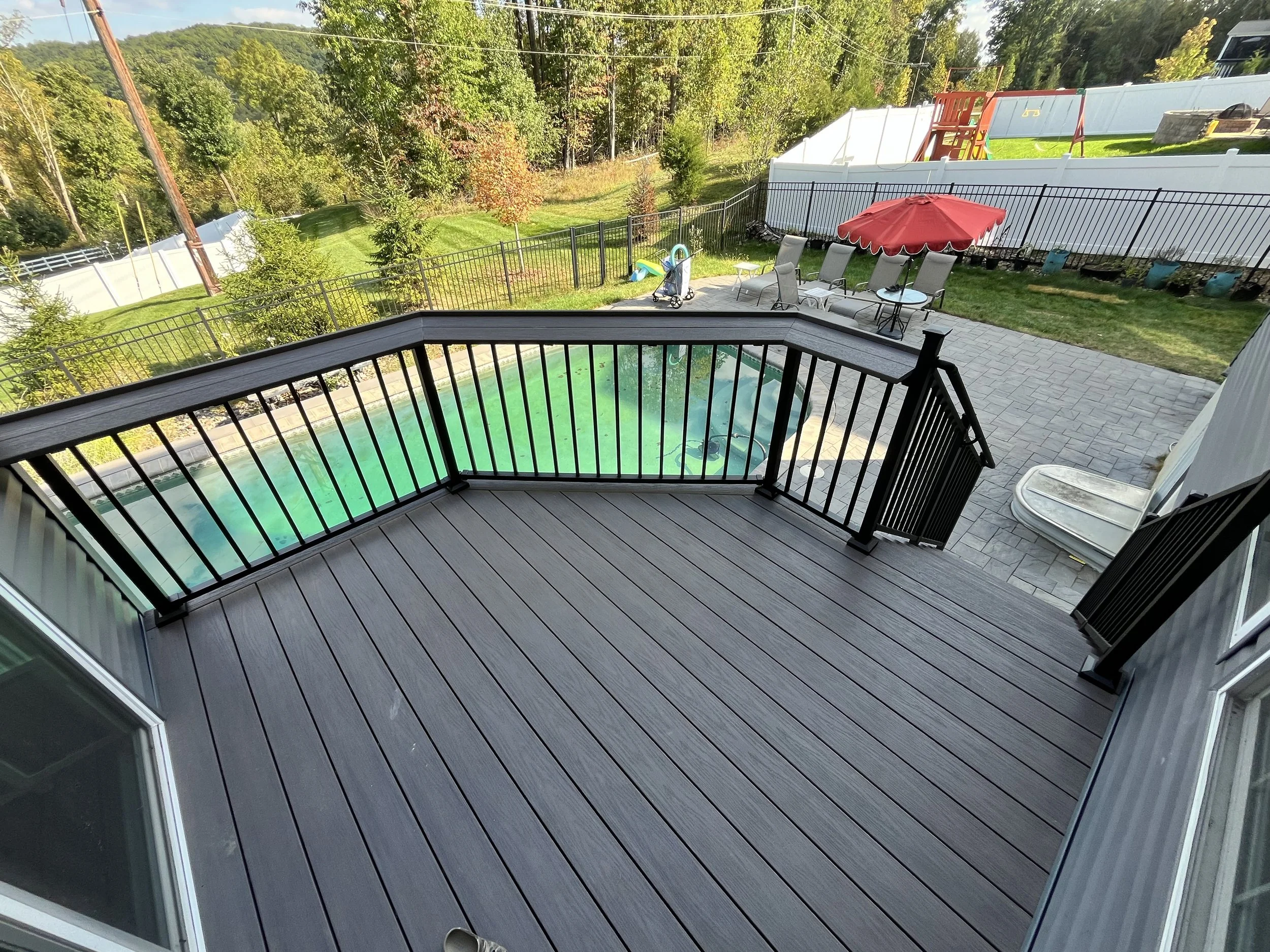 View from a balcony overlooking a backyard with a swimming pool, patio furniture, a red umbrella, and children’s play equipment, with trees and a white fence in the background.