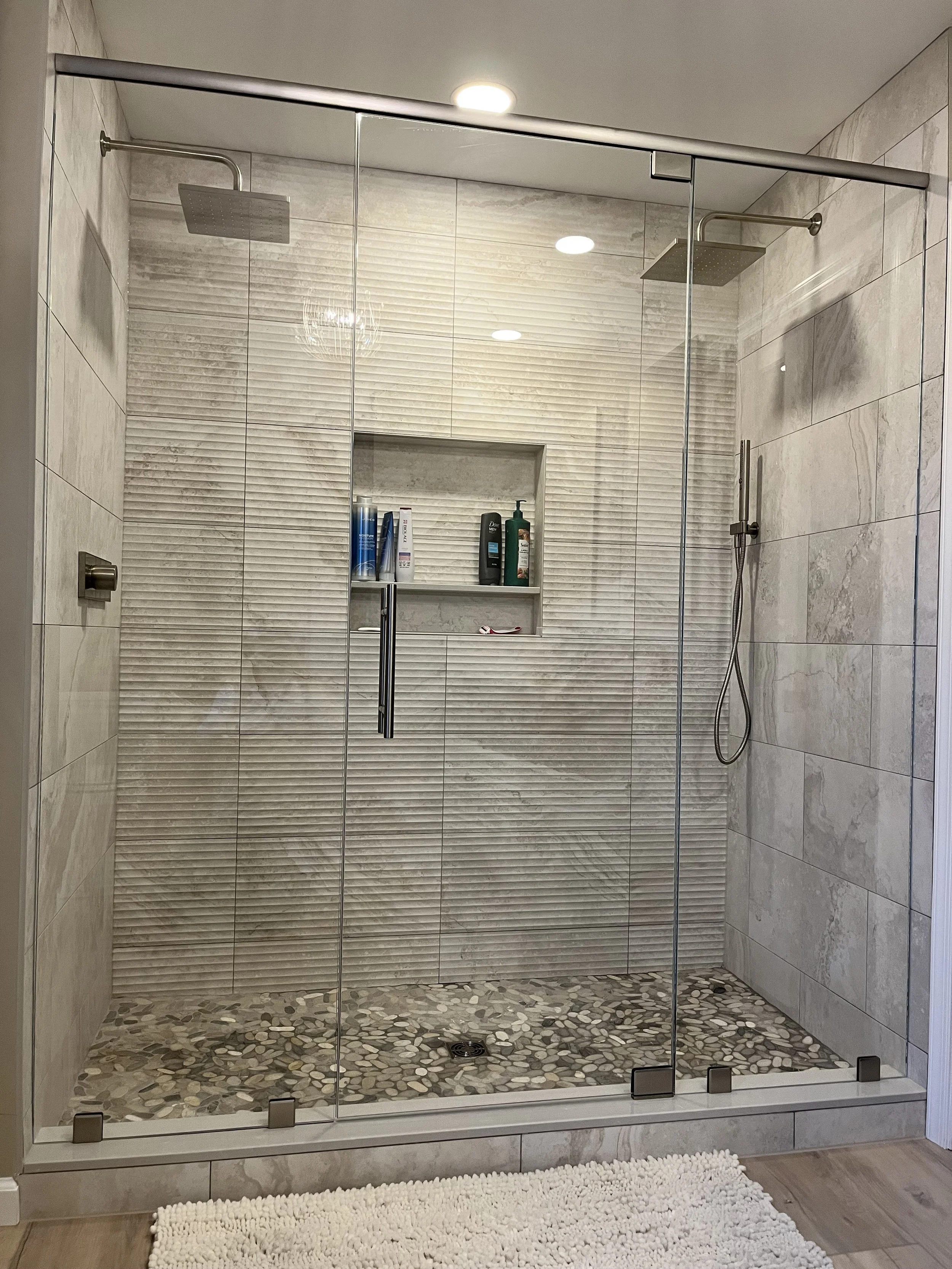 A modern walk-in shower with glass doors, two rainfall showerheads, a built-in wall niche with toiletries, pebble stone flooring, and a white textured bath mat outside the shower.