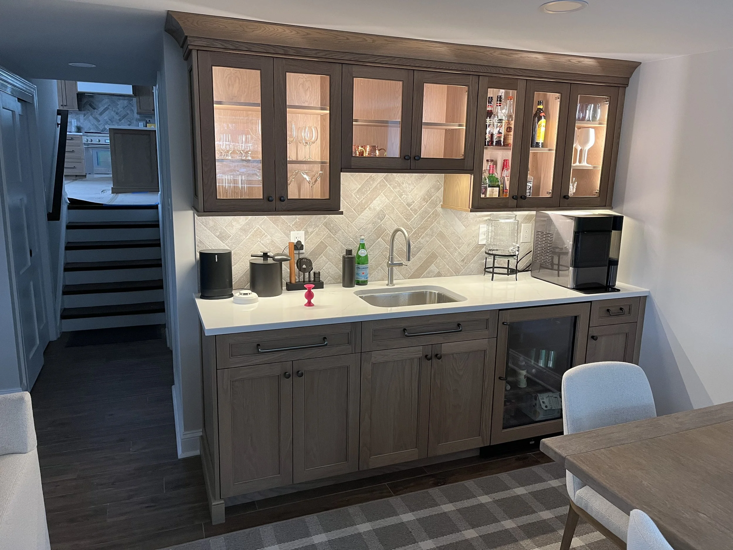 Kitchen with wooden cabinets, a white countertop, a sink, a coffee machine, and a water bottle, with a visible staircase and kitchen appliances in the background.
