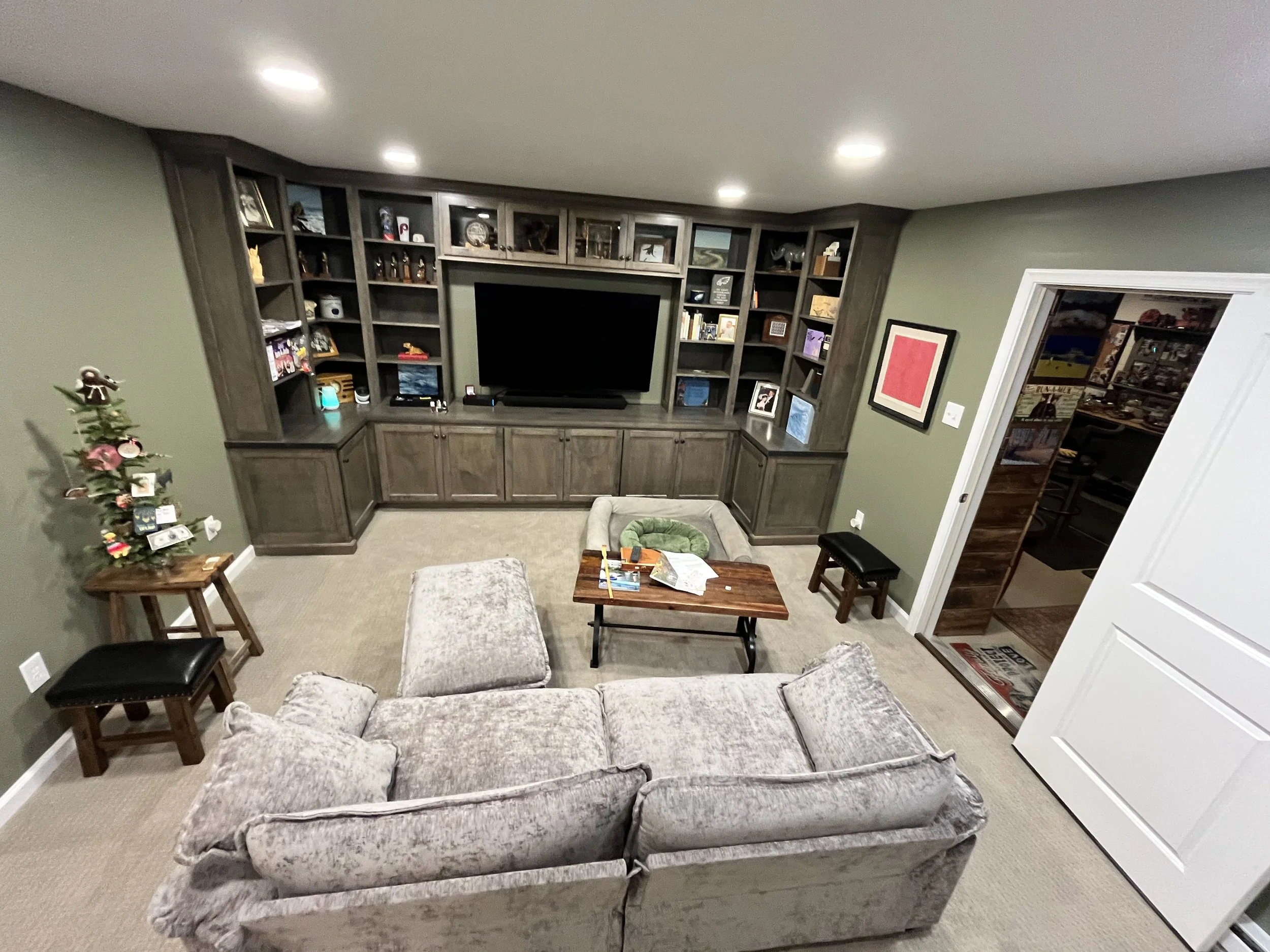 Living room with a beige velvet sofa, a wooden coffee table, and a large dark wood entertainment center with shelves and cabinets, green walls, carpeted floor, small decorated Christmas tree, framed artwork, and an open doorway leading to another roo
