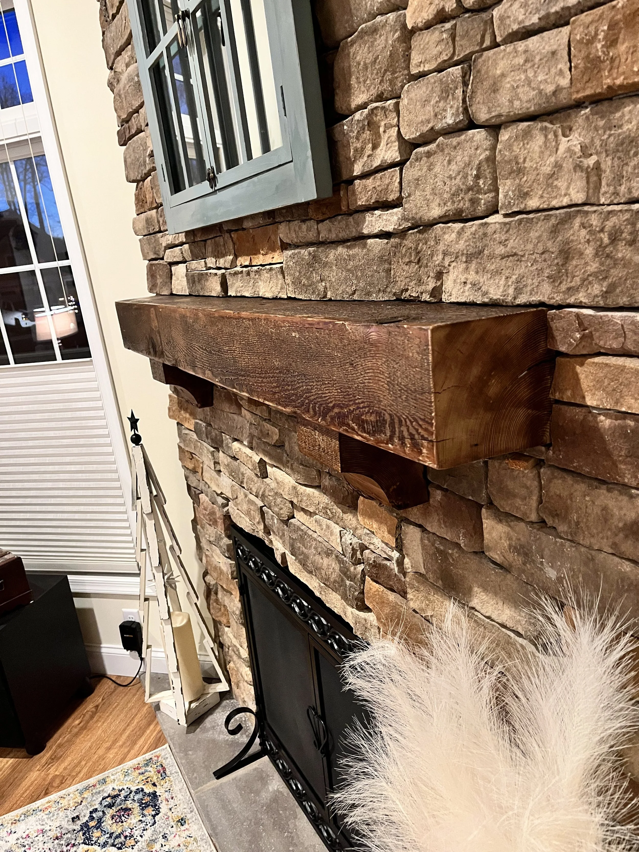 Interior view of a living room showcasing a stone fireplace with a wooden mantel, a black fireplace insert, a window with white blinds, and a glimpse of hardwood flooring, a white ladder shelf, and a colorful rug.
