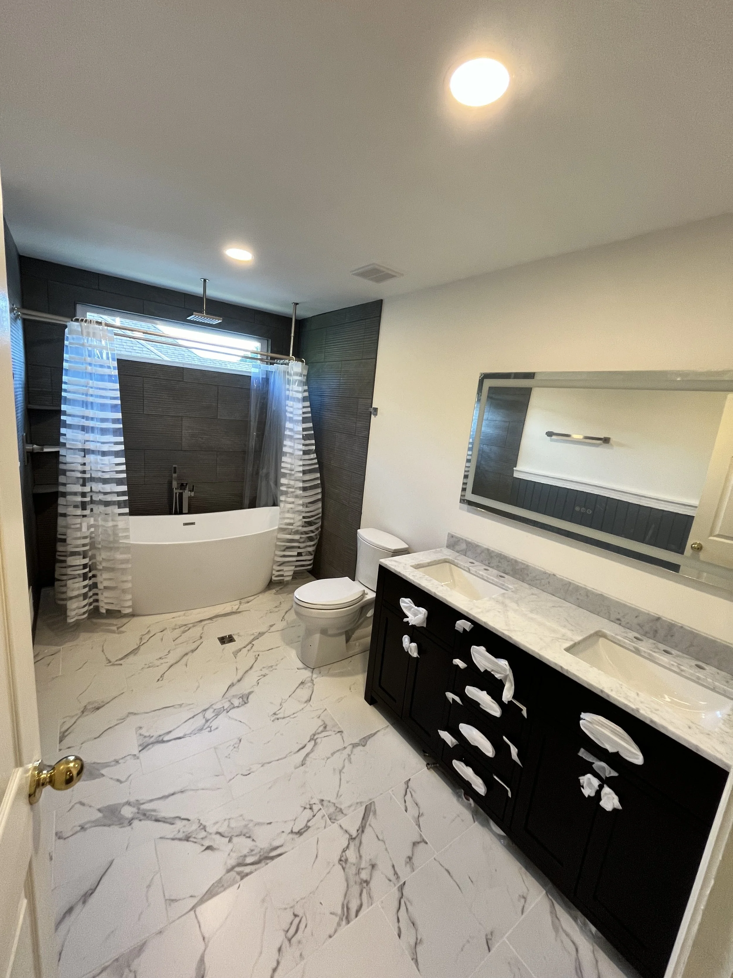 Modern bathroom with white marble floor, black vanity with two sinks, large mirror, toilet, bathtub with shower curtain, and dark accent wall near a window with blinds, ceiling lights.