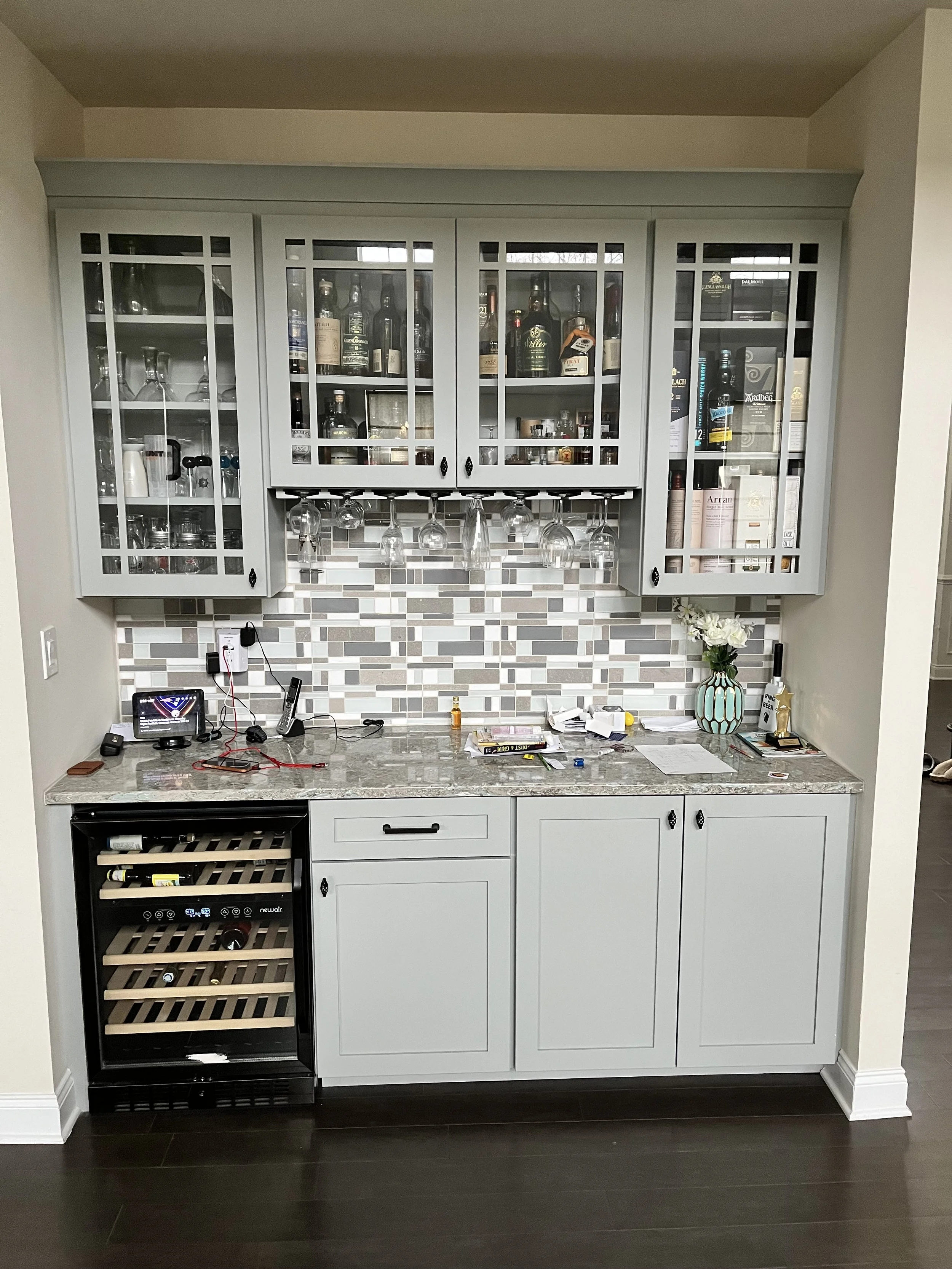 A kitchen bar area with a gray cabinet and glassware hanging underneath. The countertop has miscellaneous items, a wine cooler, a vase with flowers, and various small objects. The backsplash has multi-colored tiles.