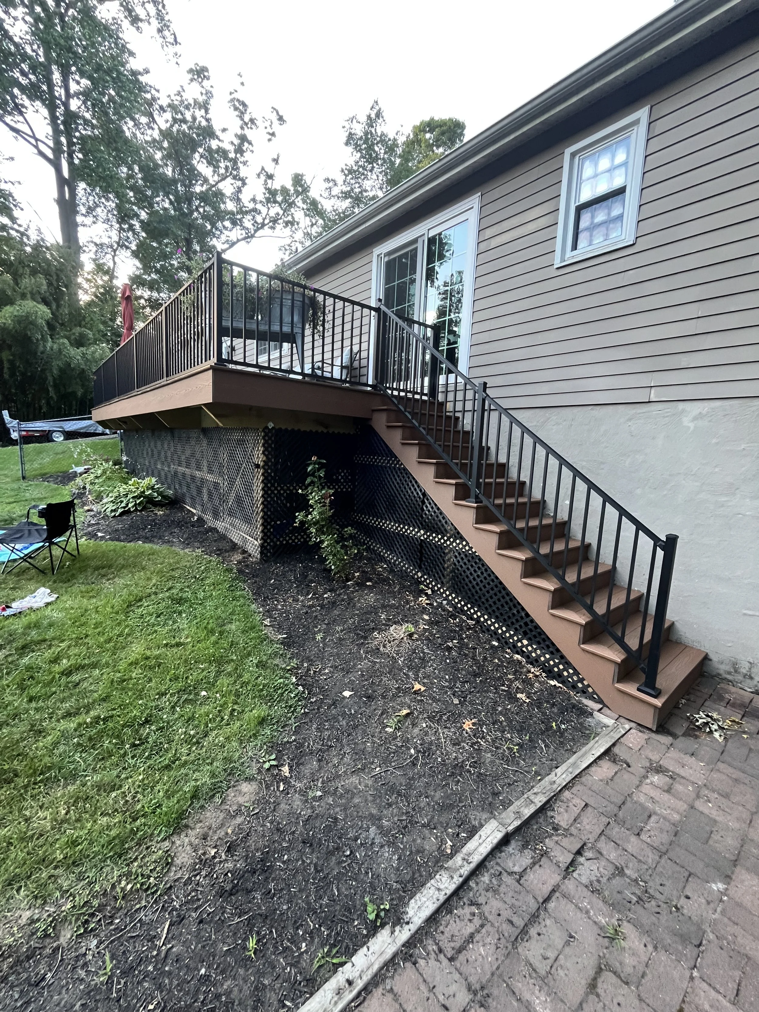 Backyard with a wooden deck and stairs attached to a house with beige siding. The yard has grass, some plants, and exposed soil. There are trees in the background and a parked car on the driveway. A garden hose is on the ground near the house.