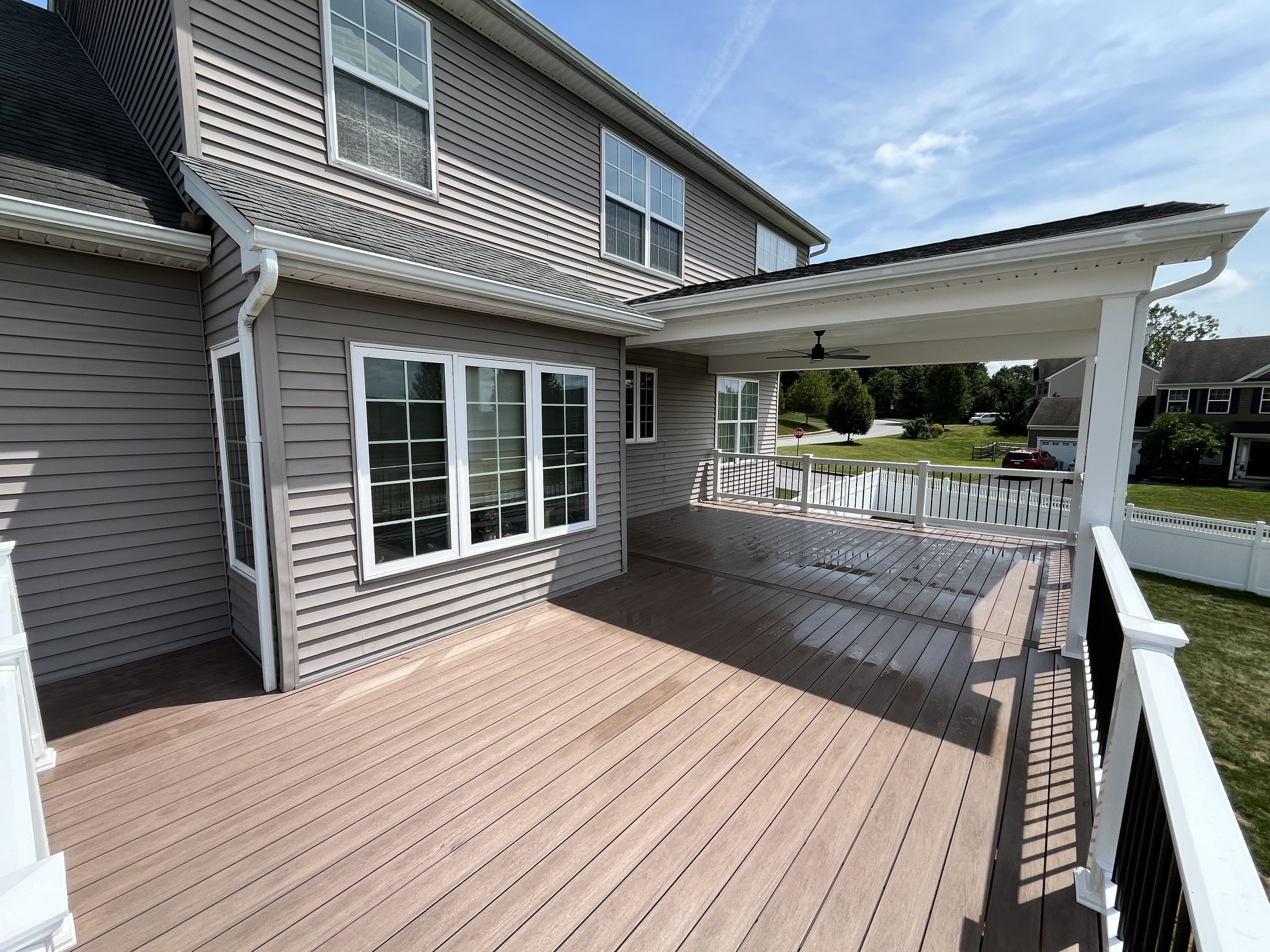 Backyard wooden deck with white railing, attached to a beige house with large windows, under a bright sky.