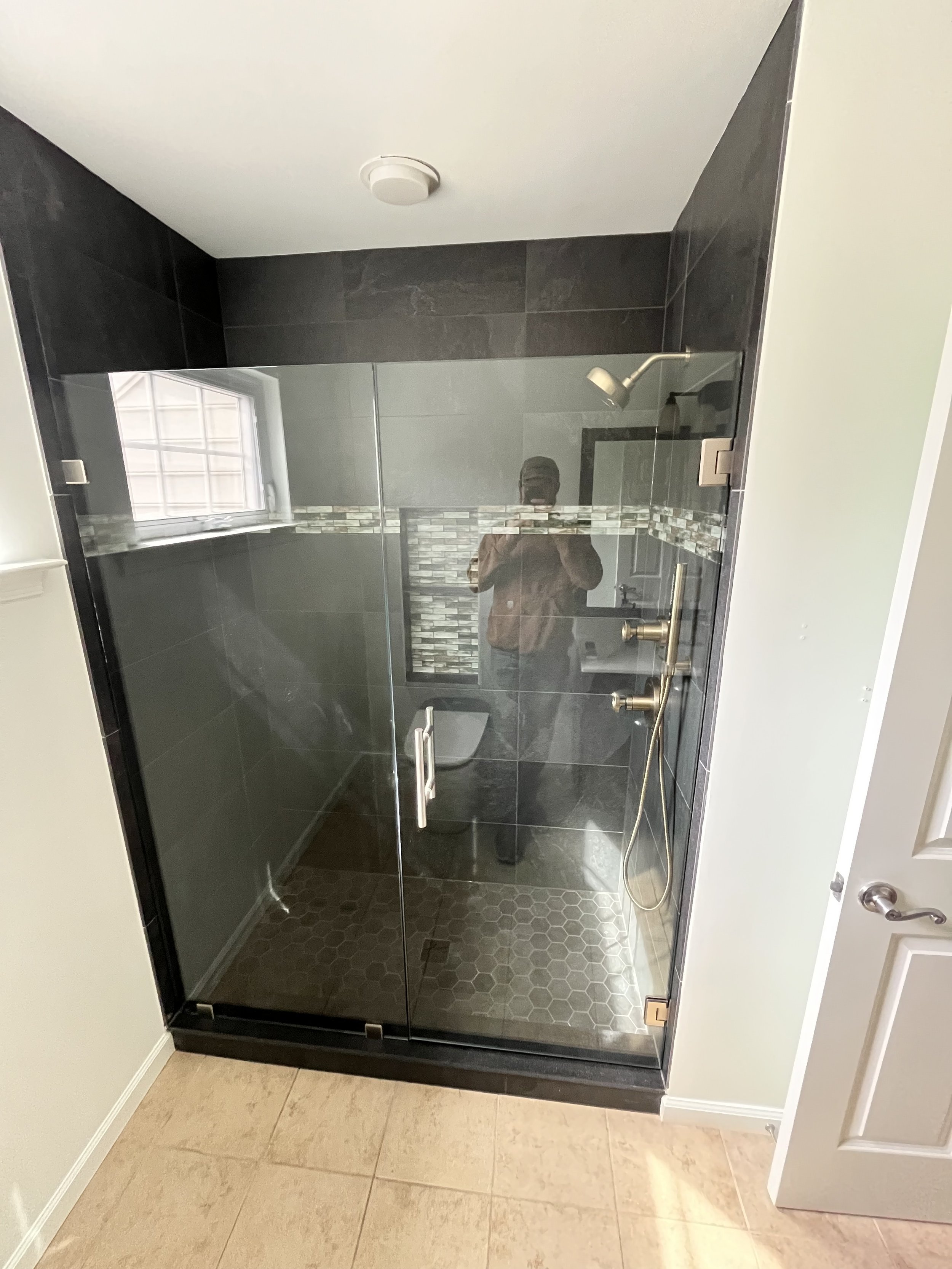 A modern shower with clear glass door, black tile walls, and hexagon floor tiles, with a person taking a photo in the reflection.