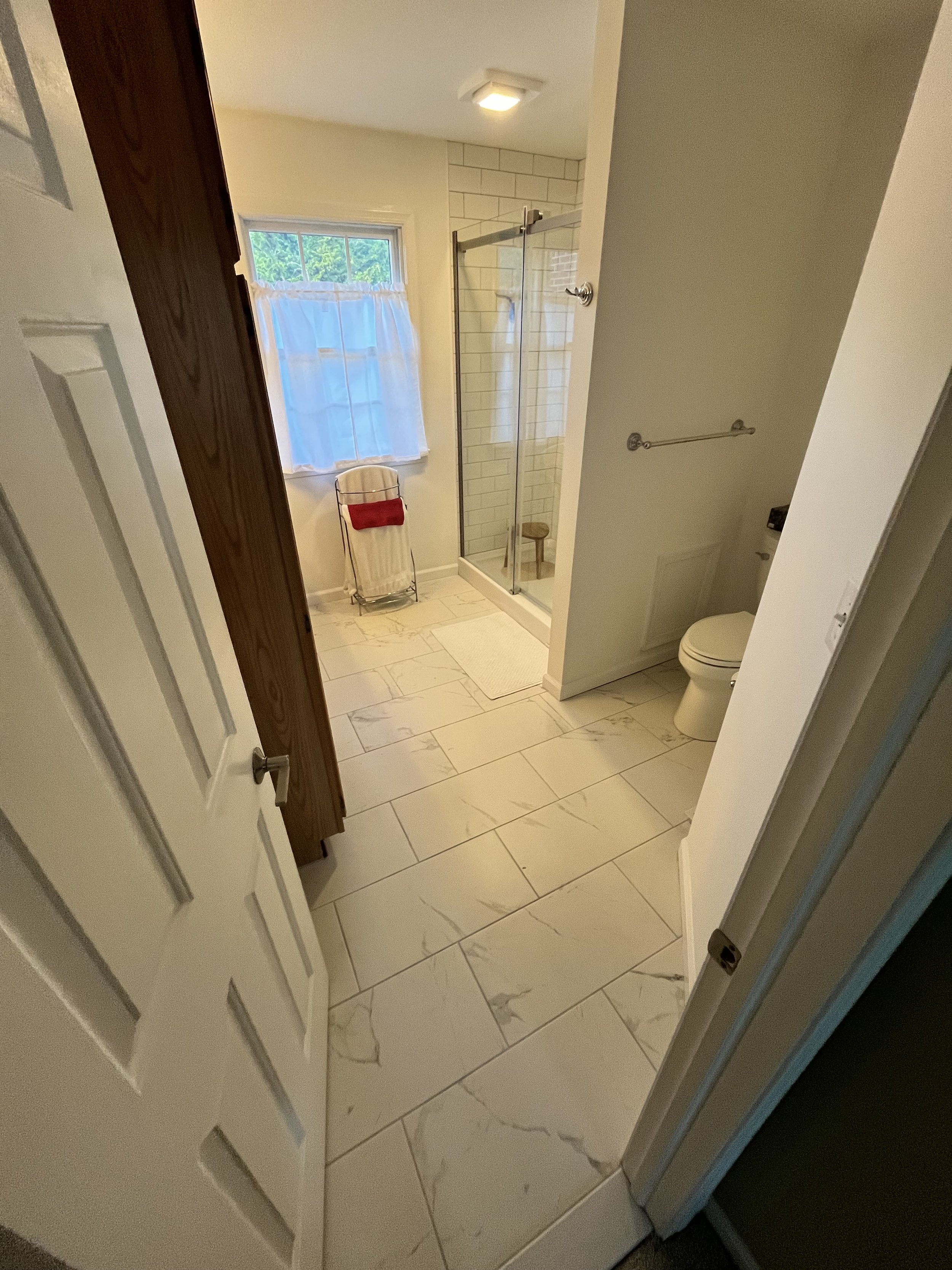 View of a small bathroom with marble tile floors, a glass shower enclosure, a window with a white curtain, a toilet, and a chair with a red towel on it.