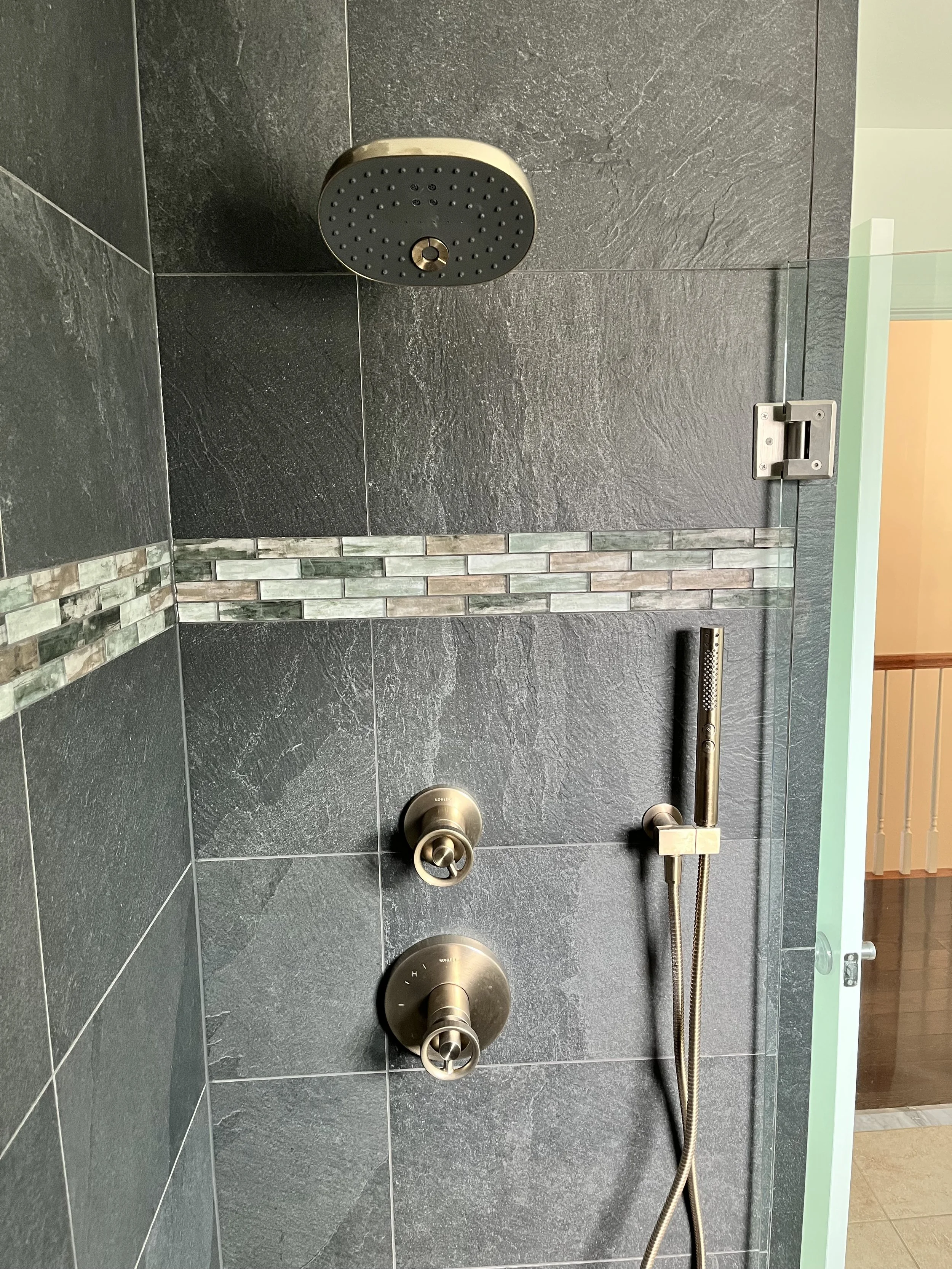 View of a modern shower with dark gray tiles, a round showerhead, a chrome handle, and a mosaic tile strip in the wall. The shower controls are visible, and part of the bathroom door and hallway are seen in the background.
