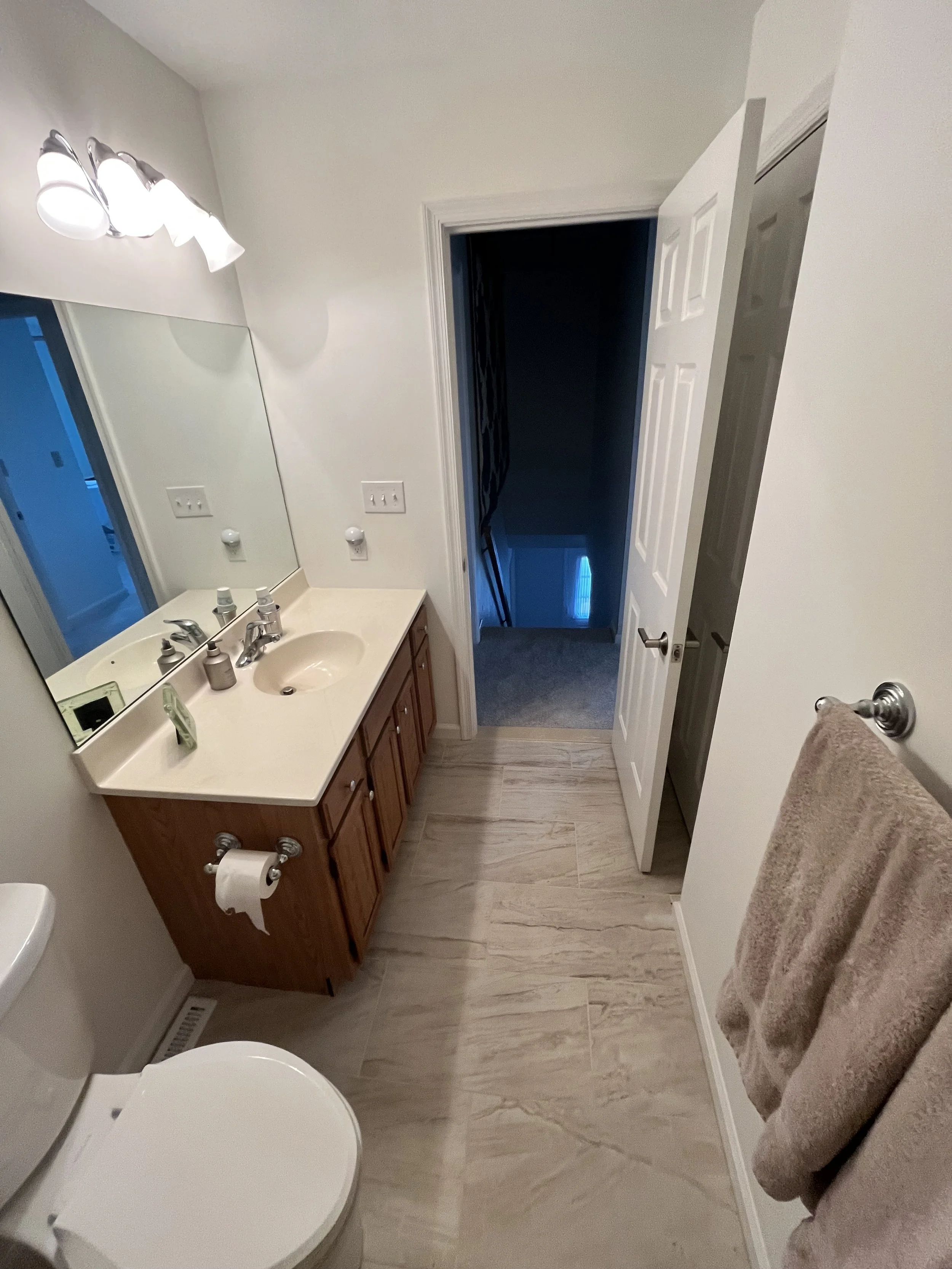 A small bathroom with a wooden vanity, white countertop, and a mirror. There is a toilet with the lid open, a towel on a rack, and a door leading to a staircase. The floor has beige tiles, and there is a light fixture above the mirror.