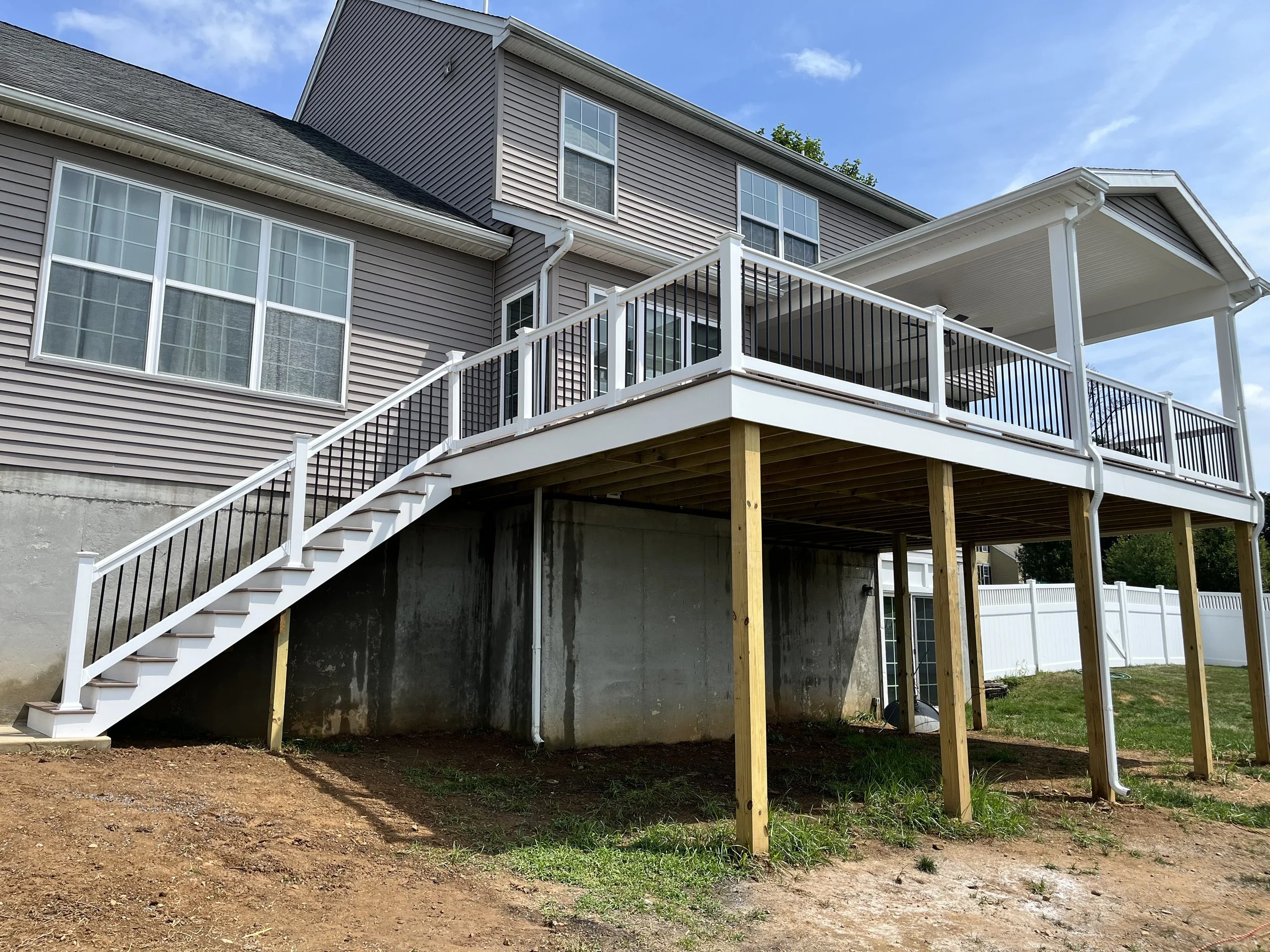 Back of a house with a raised wooden deck and stairs with black and white railing, supported by wooden posts, and a concrete foundation wall underneath, with grass and dirt ground in the foreground and a partly cloudy blue sky above.