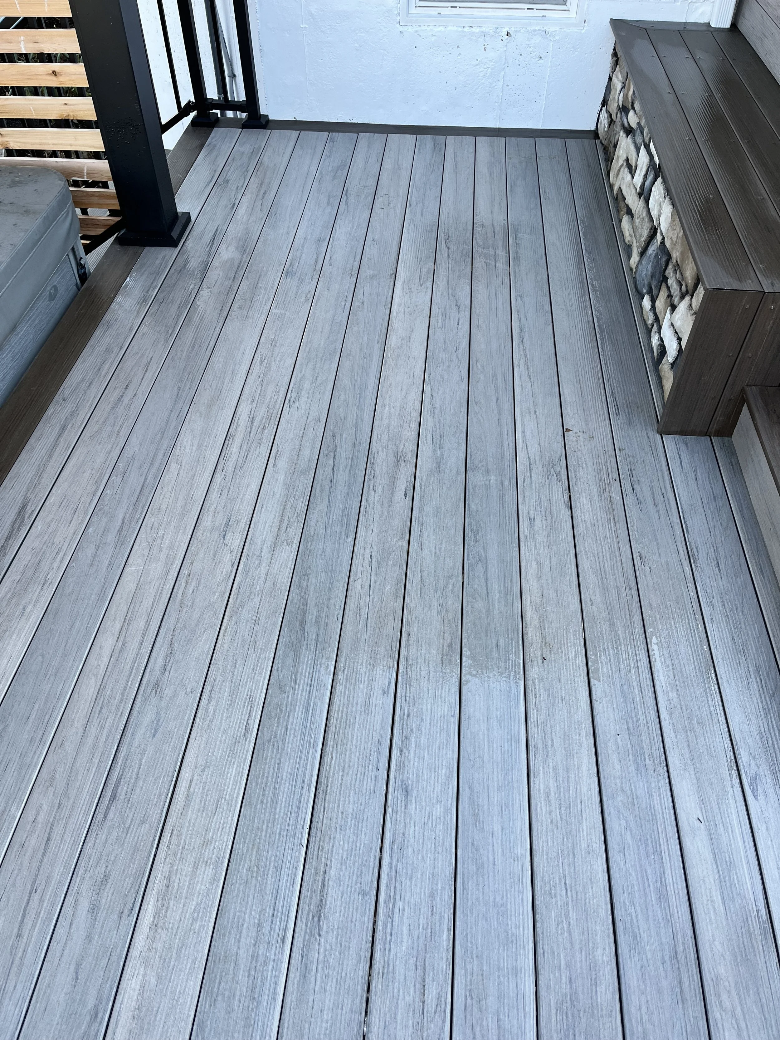 A wooden deck with grayish boards and a dark brown wooden storage bench on the right side. There is a black railing post at the corner with other railing about. A section of a metal air conditioning unit is visible in the bottom left corner. The back