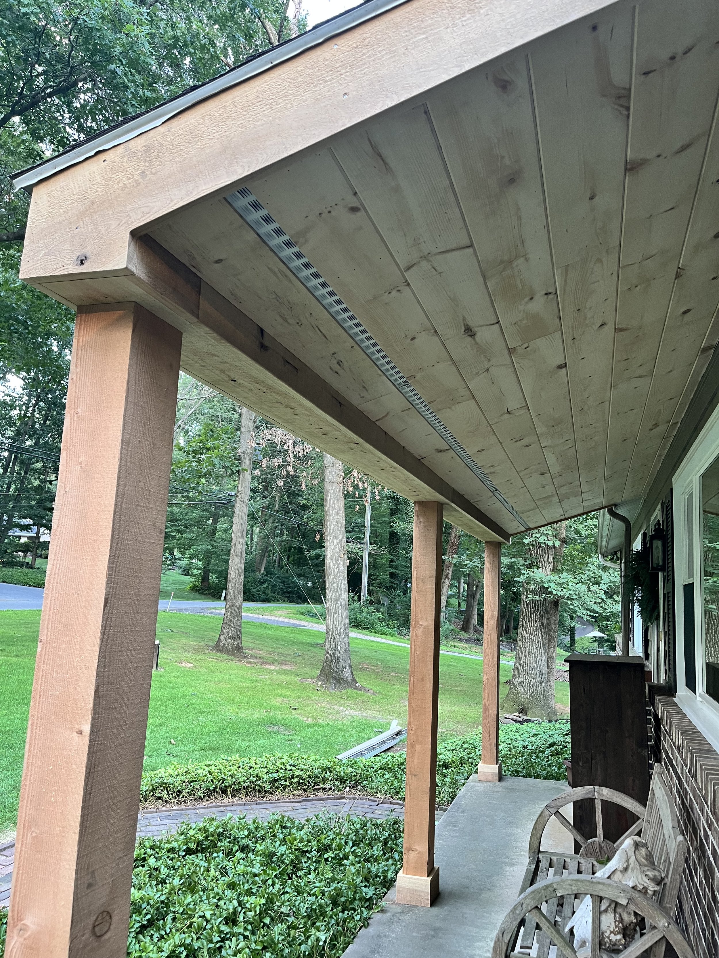 A backyard porch with a wooden roof under construction, supported by vertical posts, with a view of green grass, trees, and a small stone pathway.
