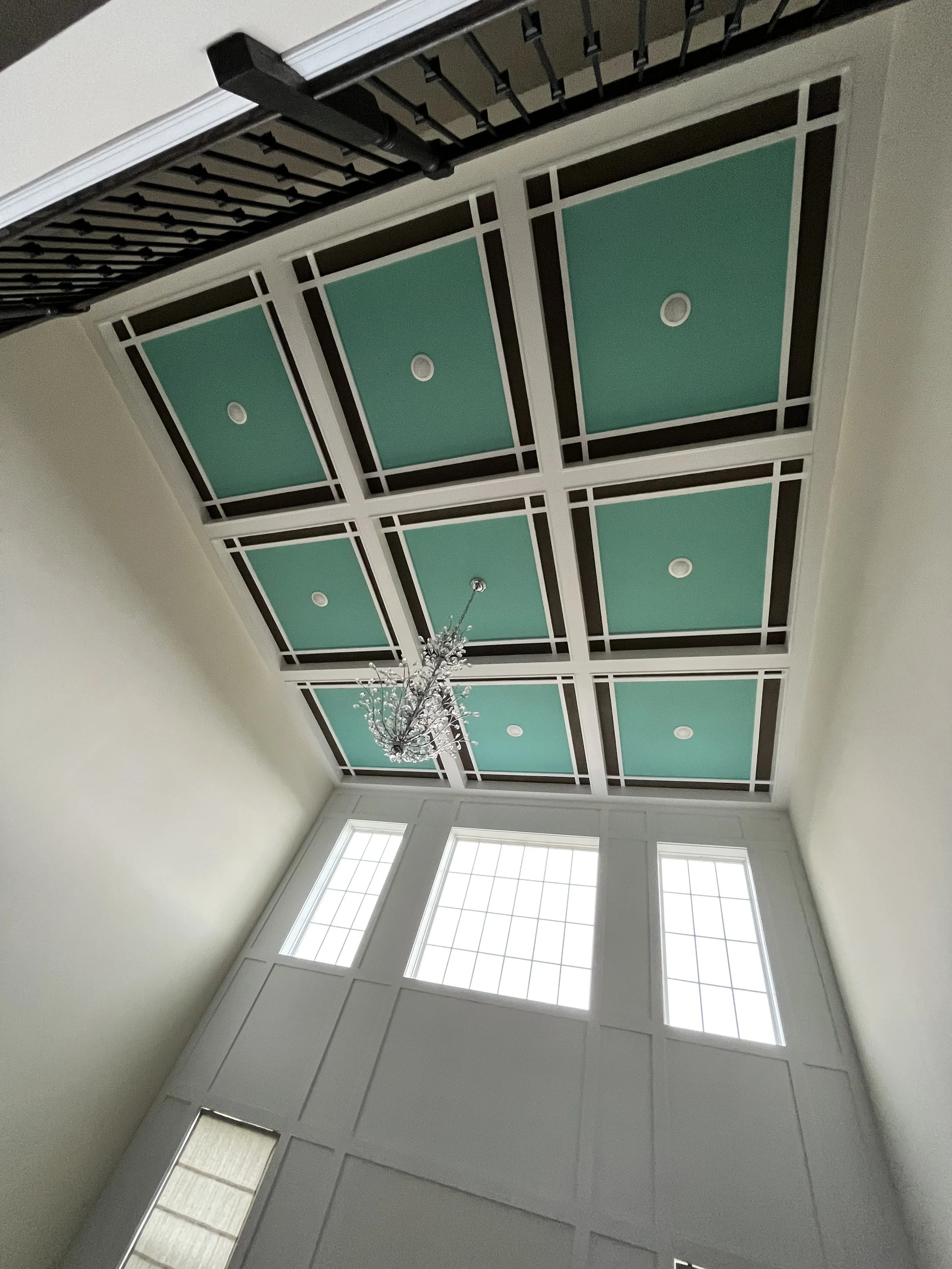 Interior view of a room with a vaulted ceiling featuring a coffered design with teal panels and white and dark brown trim, a large window, and a crystal chandelier.