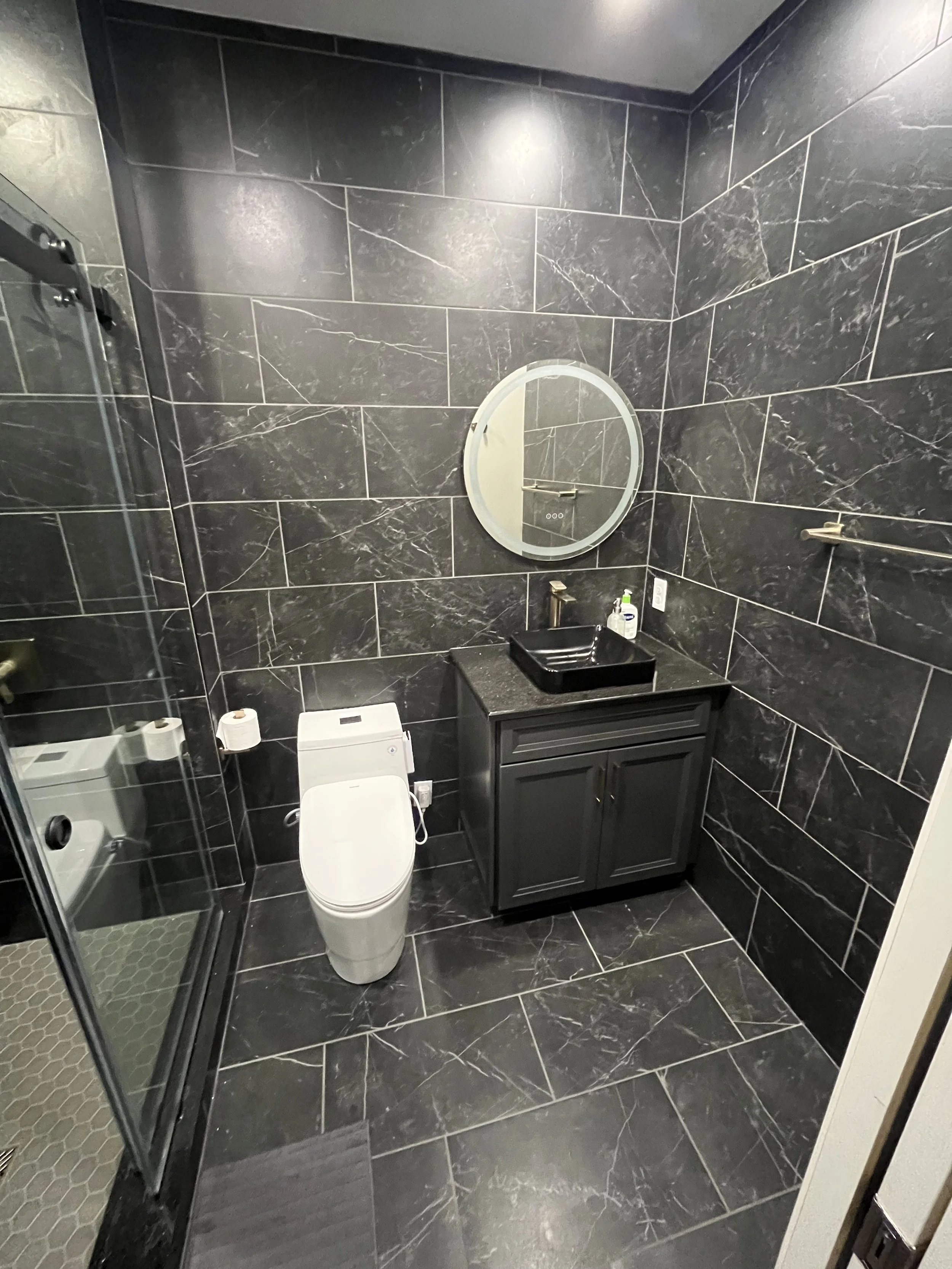 Modern black tiled bathroom with a toilet, black vanity with a black sink, round mirror, and a shower stall with a glass door.