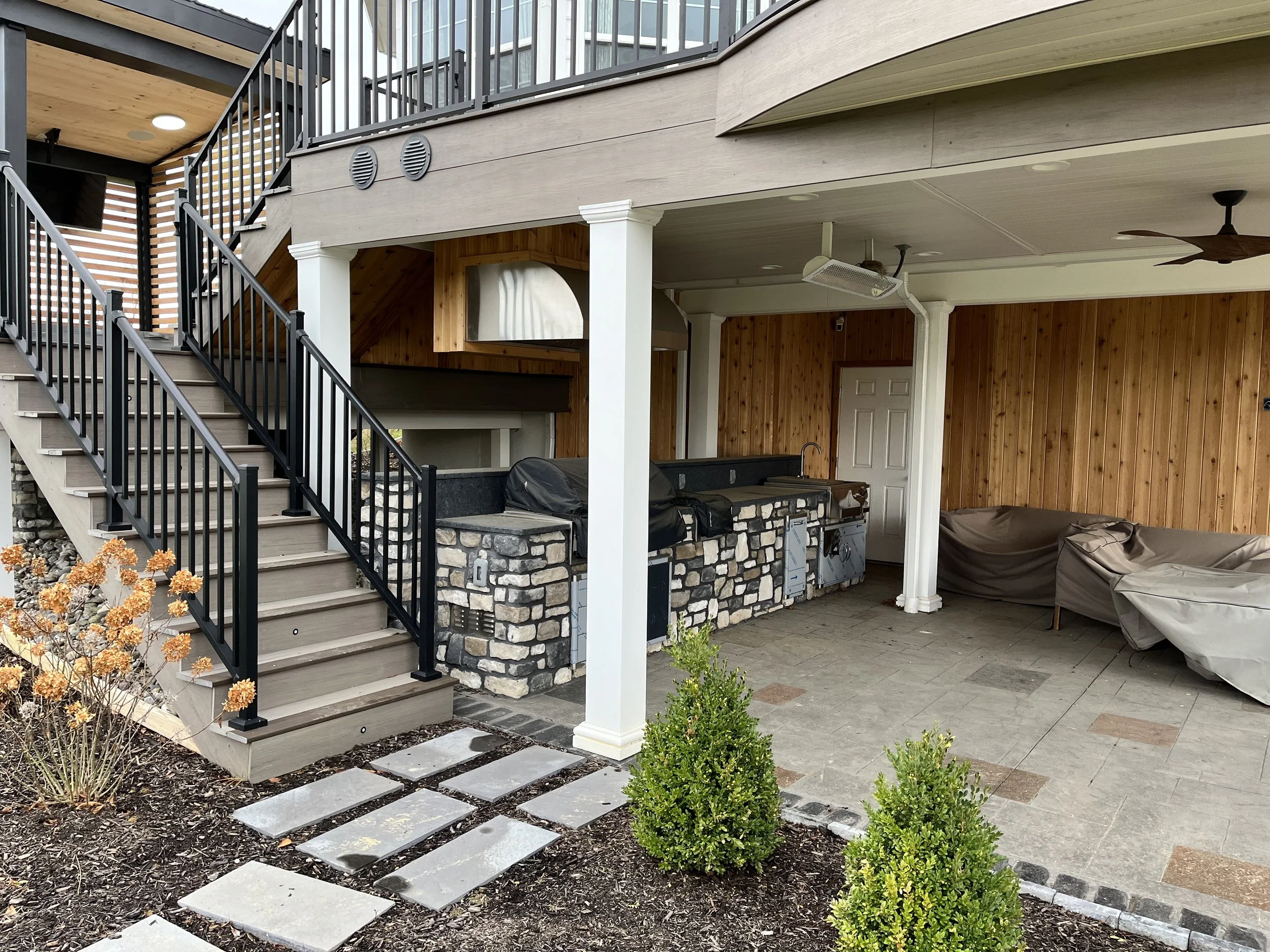 Backyard patio with wooden stairs, black metal railing, and outdoor ceiling fan. There is a covered area with a wooden ceiling, a stone fire pit, and a covered grill area with wooden paneling and a white door. potted plants and bushes are in the gard