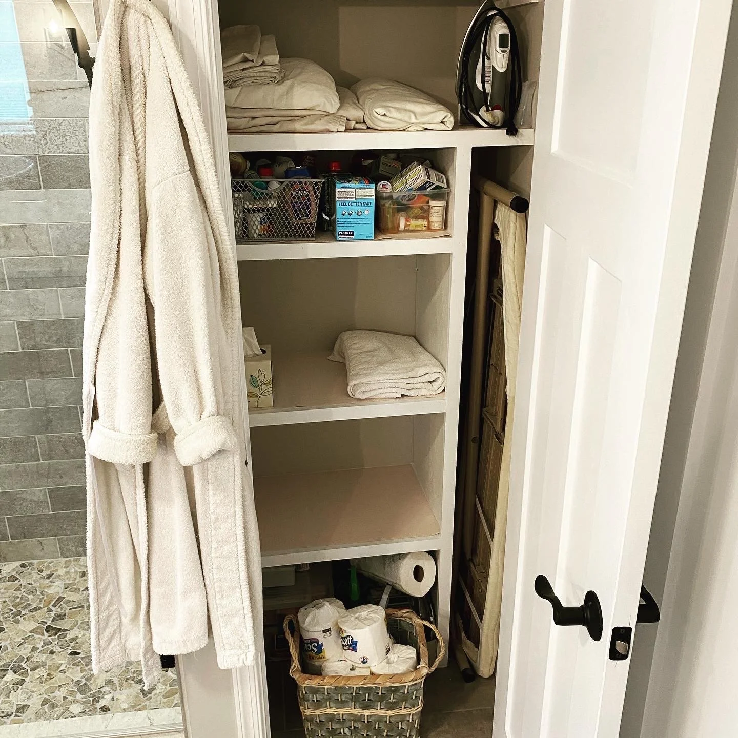 A narrow closet with white shelves containing folded towels, a basket with supplies, and boxes. A lint roller, cleaning supplies, and various household items are stored inside. A white bathrobe hangs on the outside of the closet door.