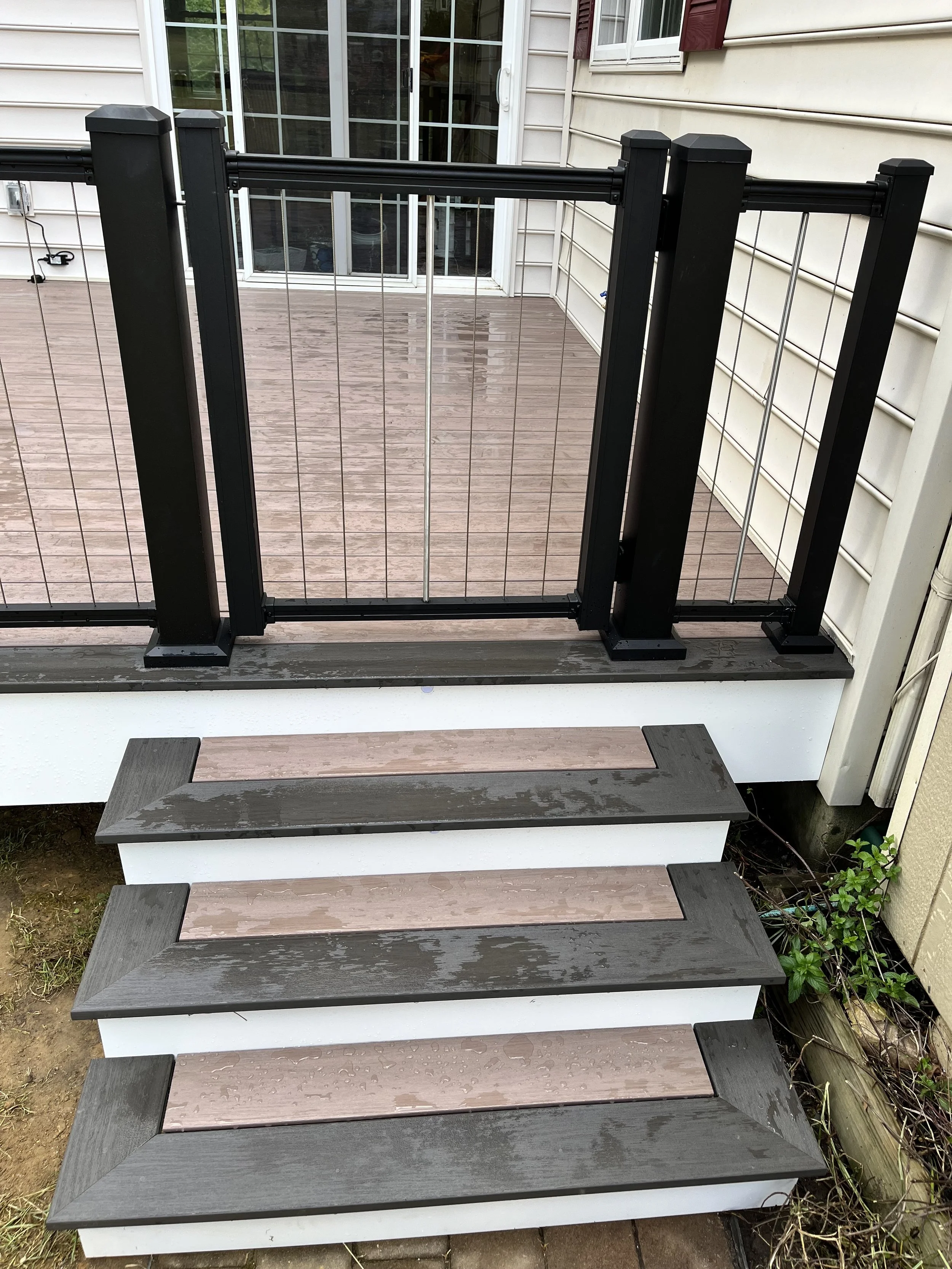 A wooden deck with a black metal pet gate blocking the stairs, with sliding glass doors and cream-colored house siding in the background.