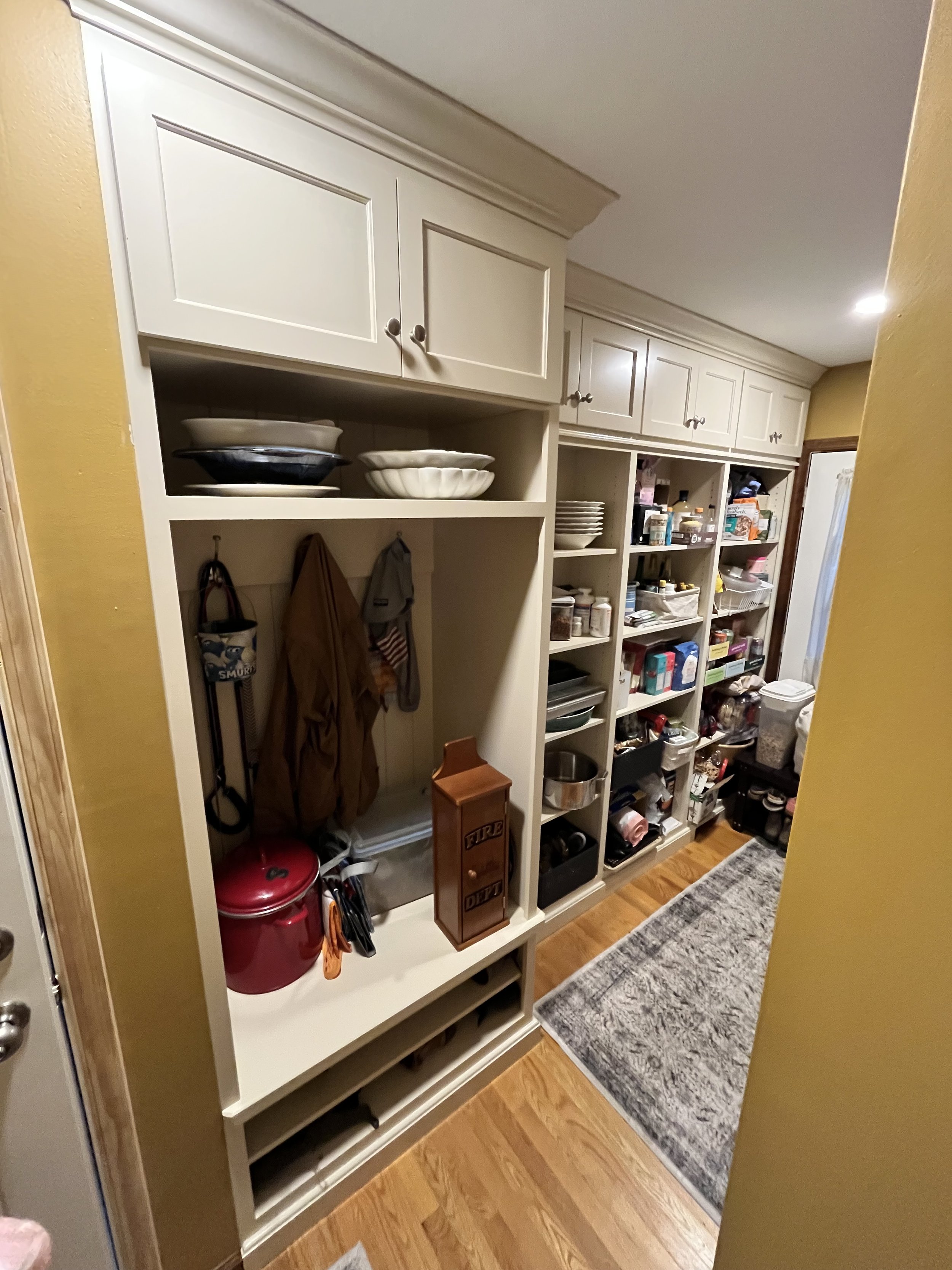 A pantry with built-in white shelves filled with dishes, containers, and various household items, with a wood floor and part of a gray rug visible.