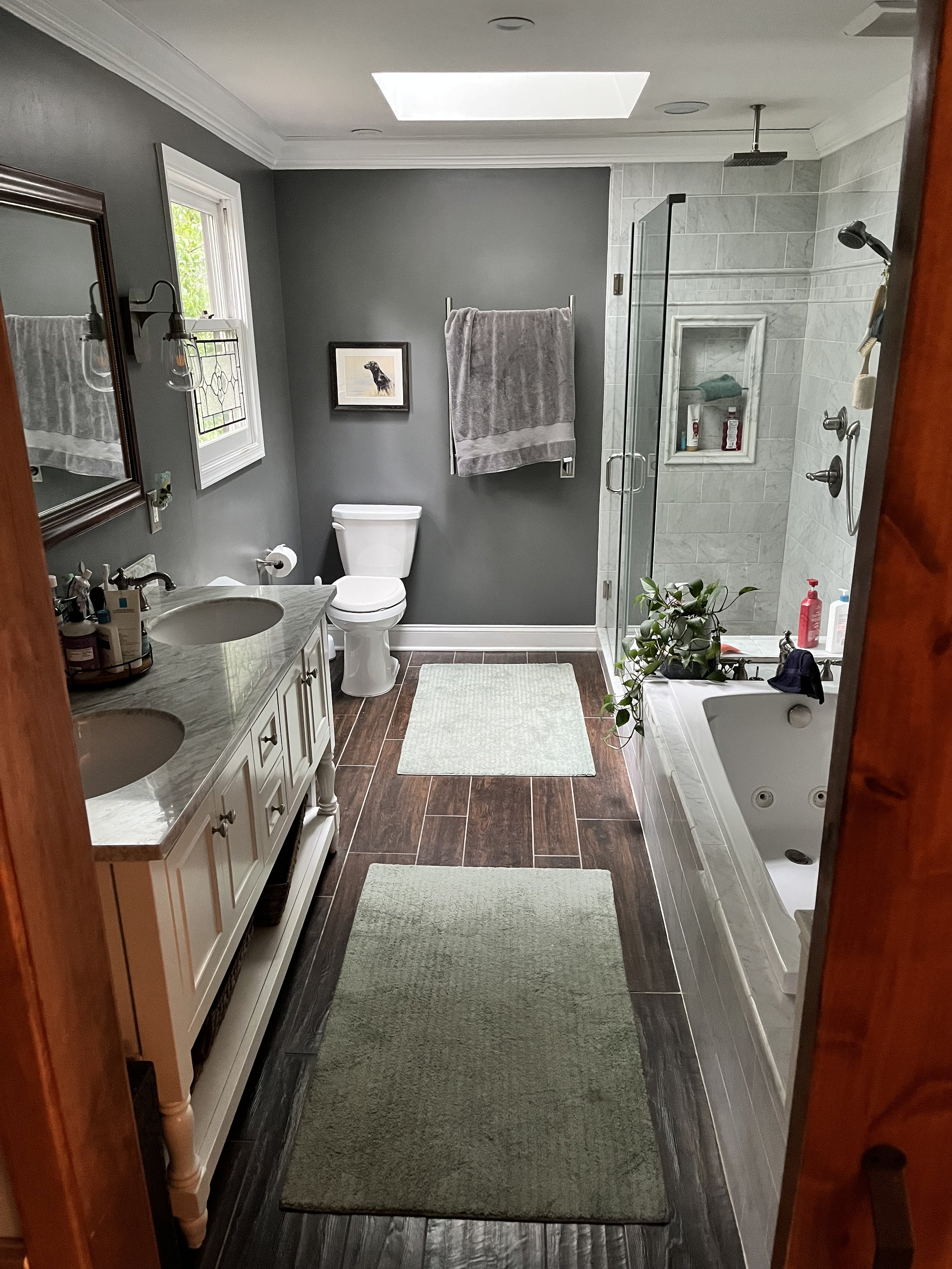 Bathroom with gray walls, double vanity with marble countertops, mirror, two sinks, towel rack with gray towel, small framed dog picture, toilet, window, skylight, shower with glass door, built-in niche, plants, bath products, small light-colored rug