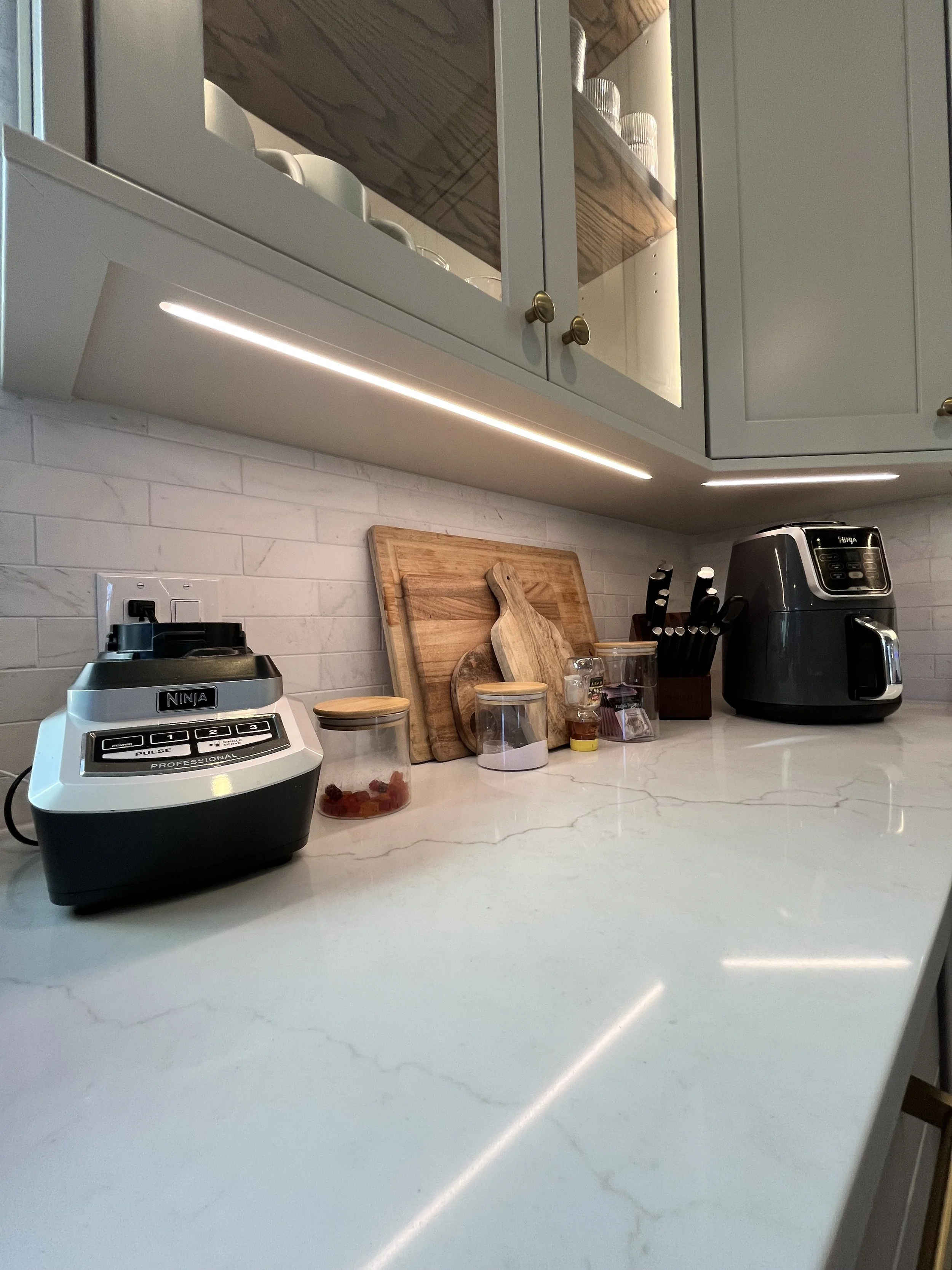 A kitchen countertop with a food processor, wooden cutting boards, glass jars, a knife block, and an air fryer.