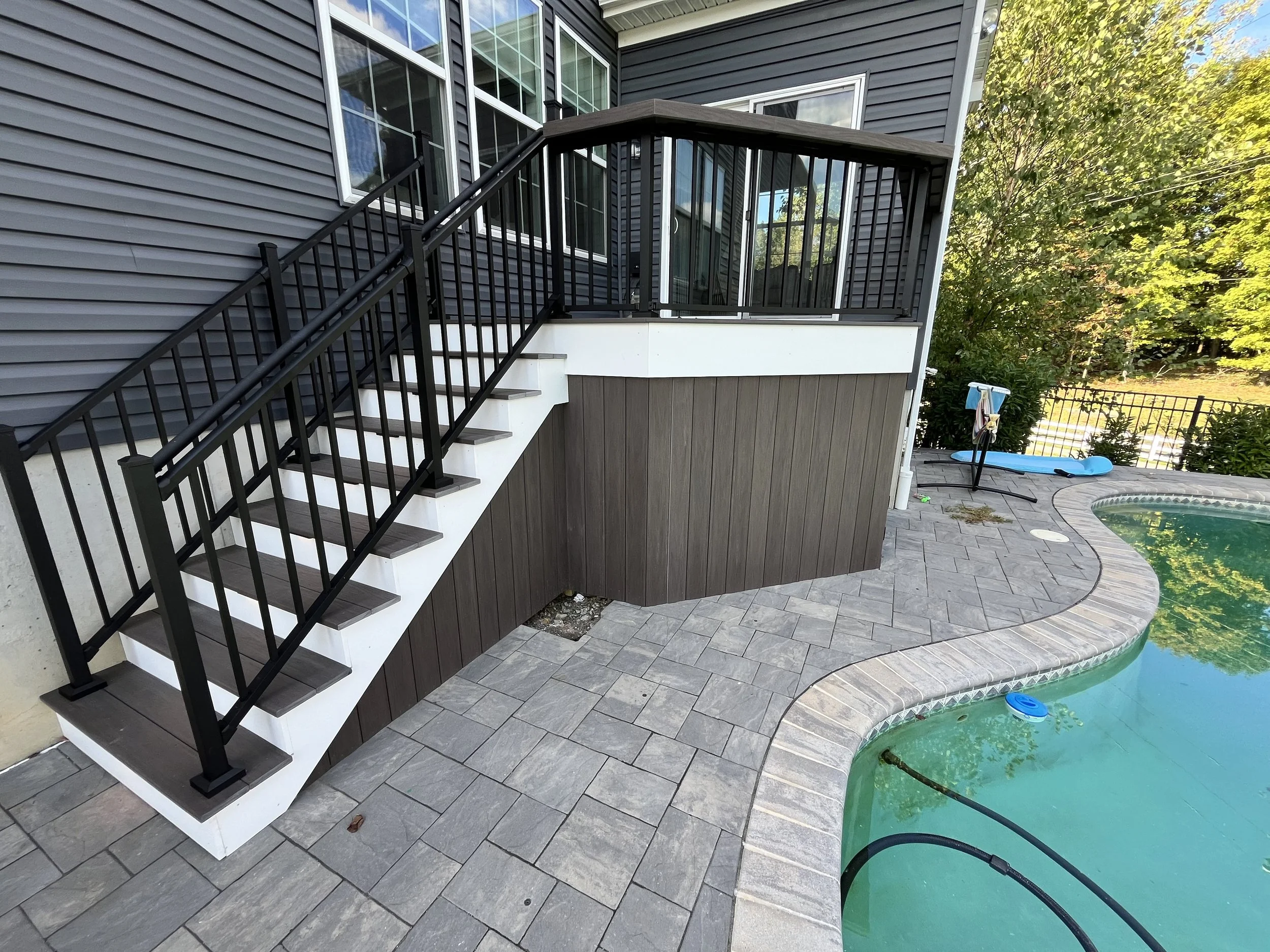 A backyard with a small pool, a patio with gray stone tiles, a black metal staircase with wood steps, and a small enclosed balcony with black railing and glass walls. Trees with green leaves are in the background.