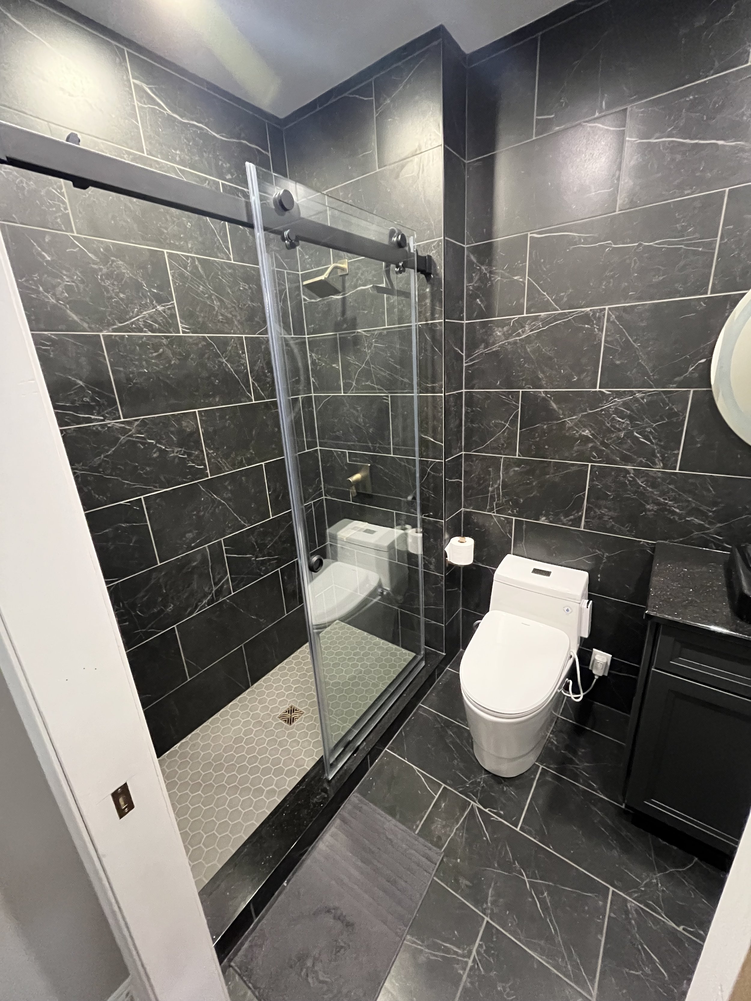 Modern bathroom with black marble tiles, a small glass shower enclosure with a sliding door, and a white toilet. There is a small black cabinet and a countertop with toiletries.