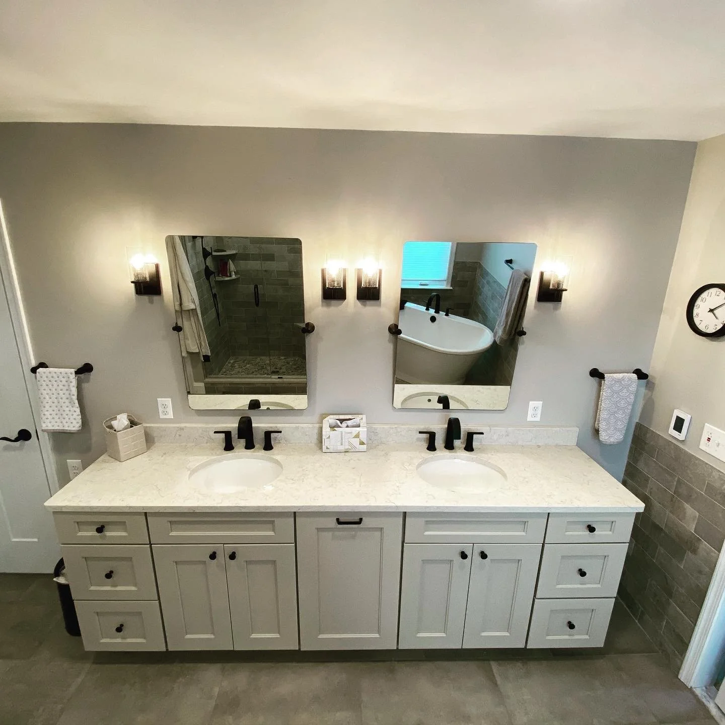 Double bathroom vanity with two sinks and two mirrors, dark fixtures, towels, tissue box, and a wall clock in the background.