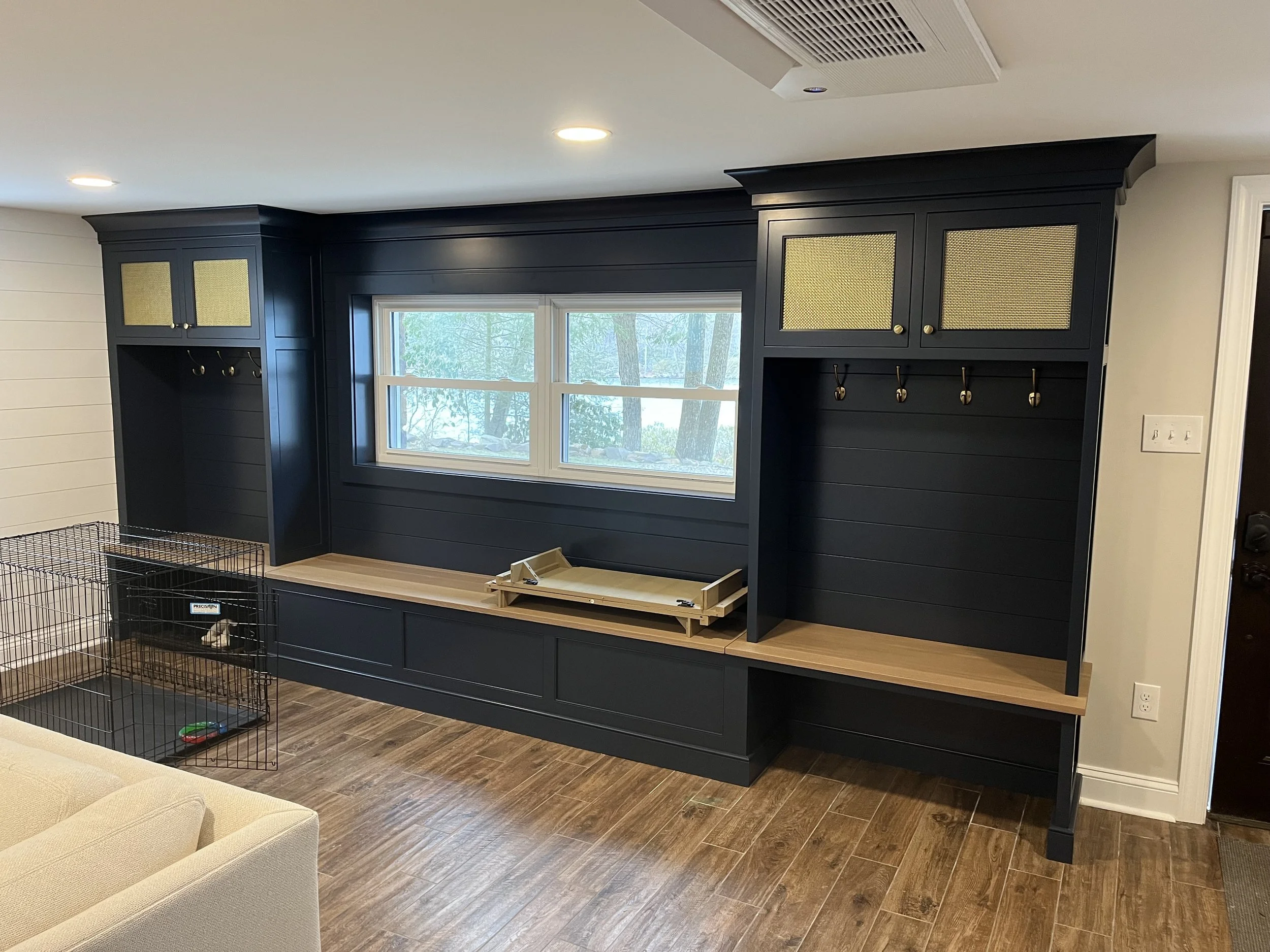 Interior of a room with a black built-in wall unit featuring gold mesh cabinet doors and hooks, a window showing trees outside, a beige chair in the foreground, a small pet crate in the corner, and hardwood flooring.