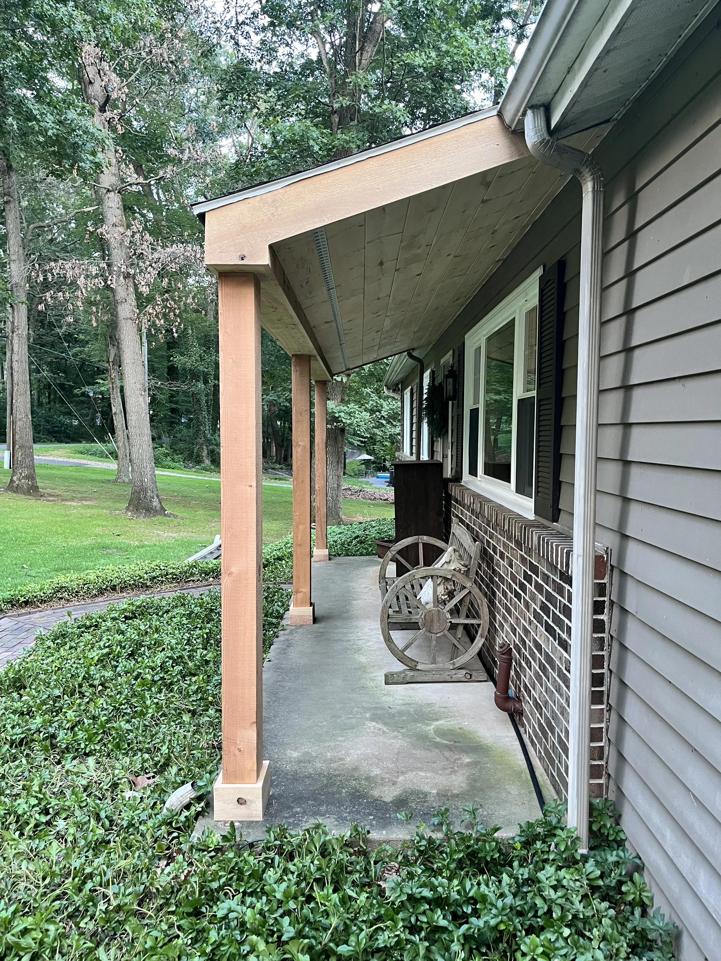 Side view of a house with a covered porch, brick wall, and window, surrounded by green bushes and trees, with construction supports for a porch extension.