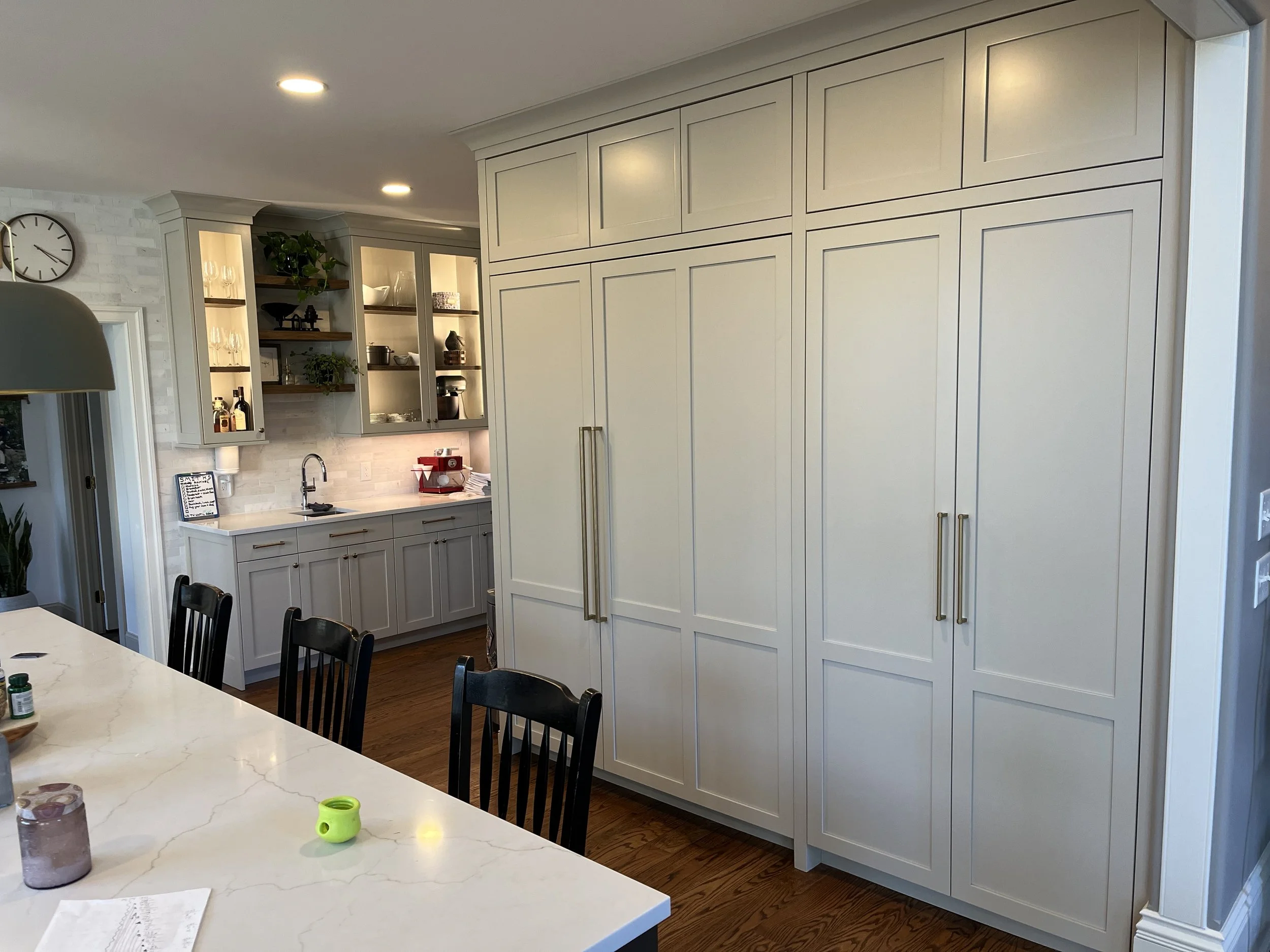 Kitchen with white cabinets, a countertop with a sink, open shelves with glassware and decorative items, a large clock on the wall, and a dining table with chairs. There are some drinks and papers on the table.