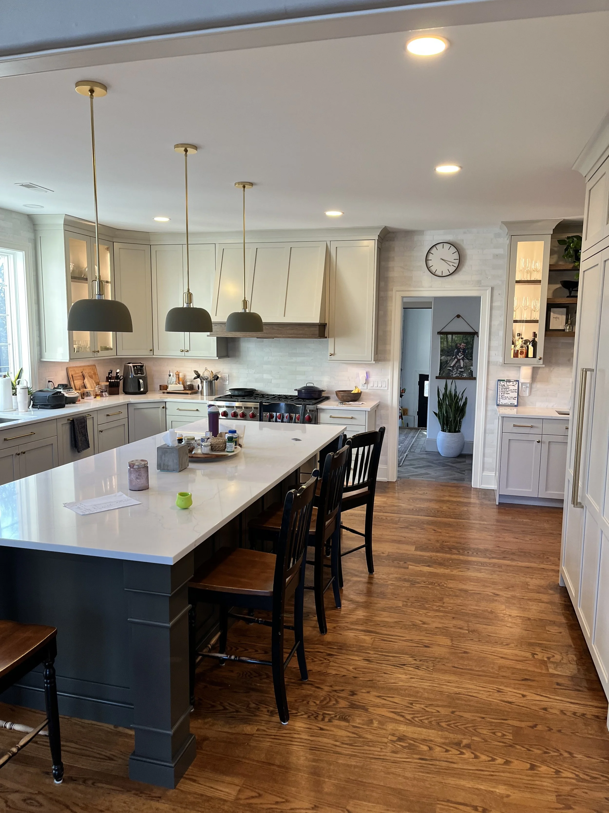Kitchen with white cabinets, a large white island with black chairs, pendant lights, and hardwood flooring. There are small kitchen appliances on the counter and a doorway leading to another room with a plant and wall decor.