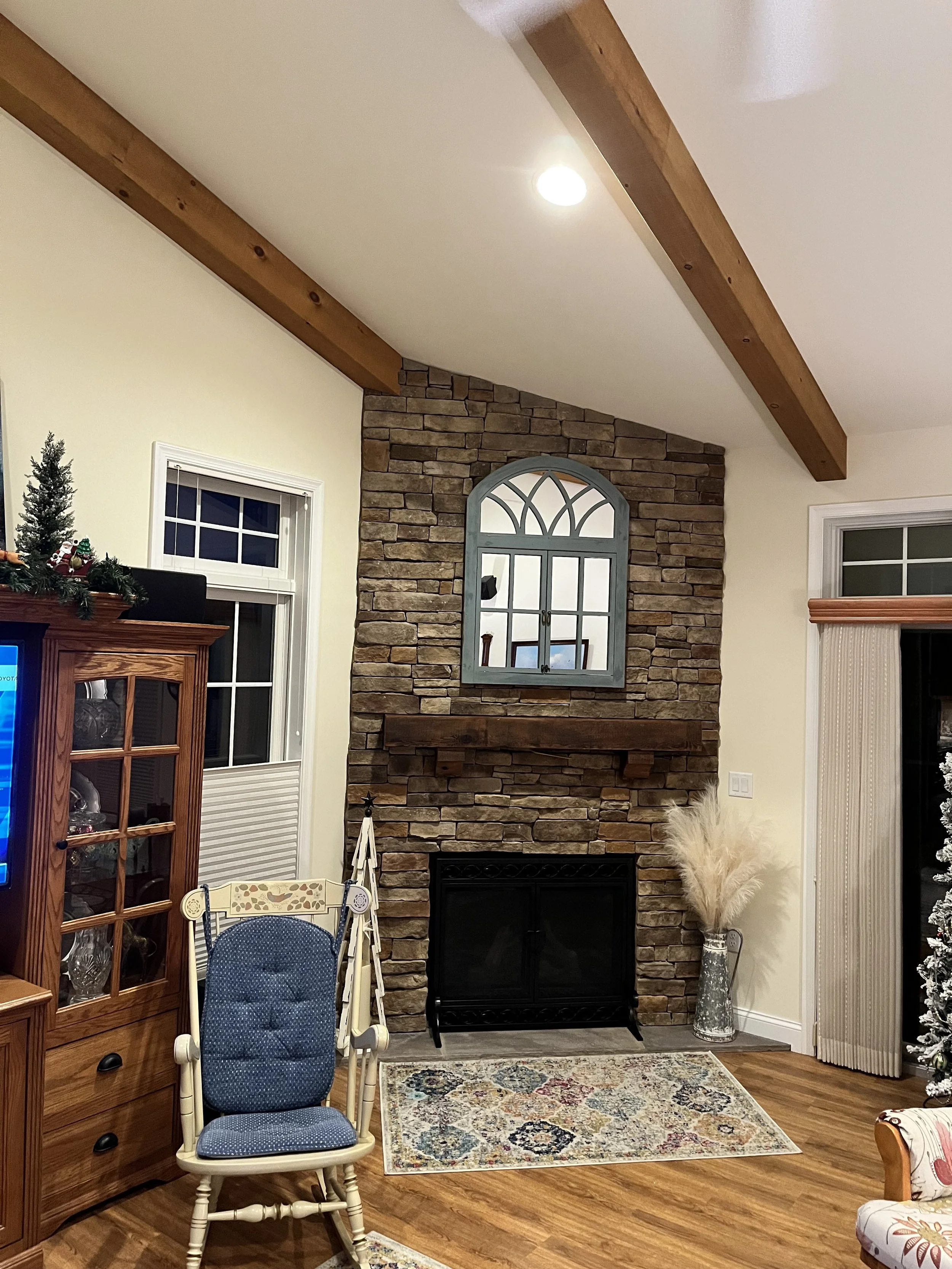 Interior of a living room with a stone fireplace, a decorative mirror above the fireplace, wooden beams on the ceiling, a window with blinds, a vase with pampas grass, and furniture including chairs and a cabinet.