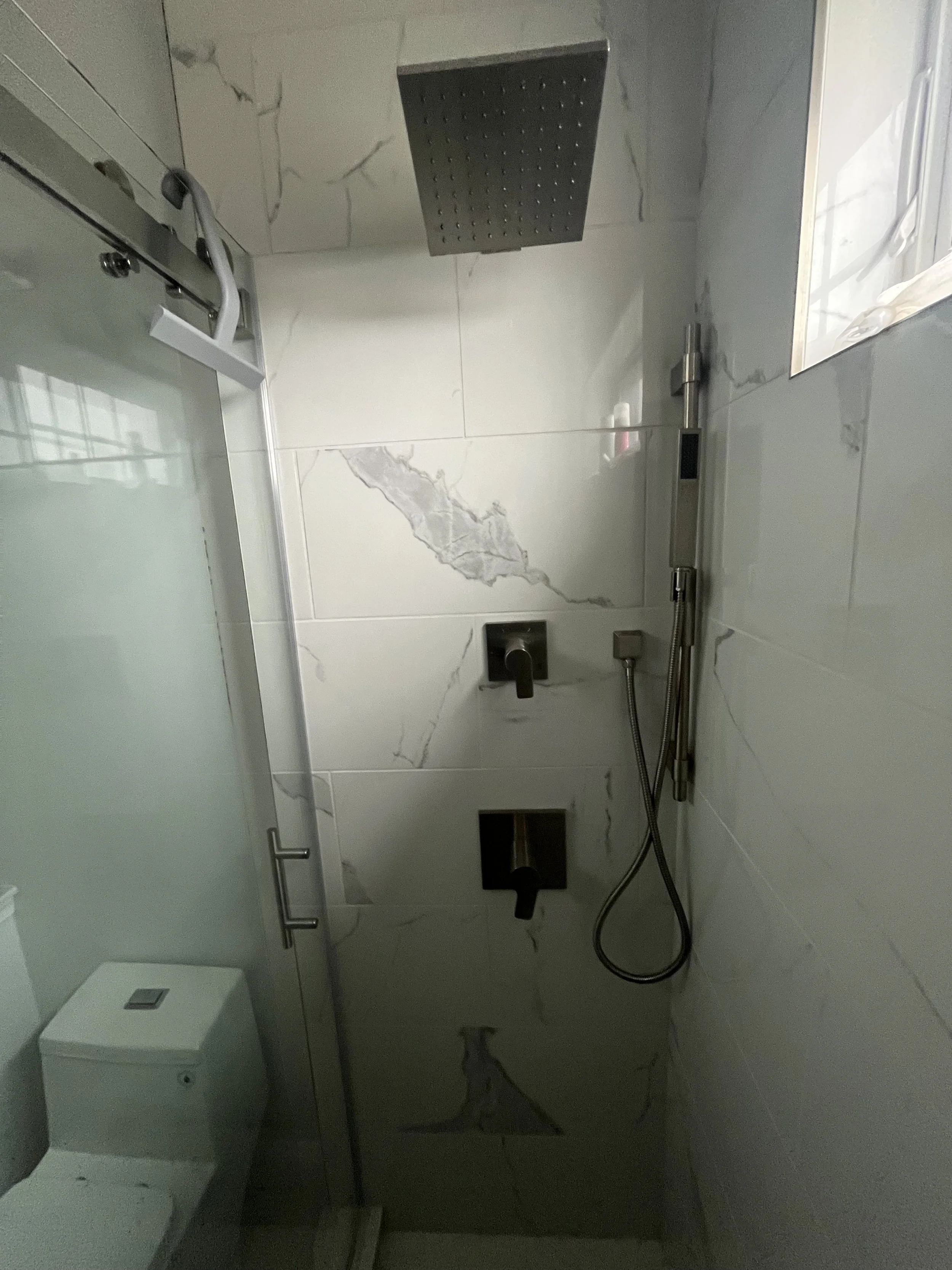 A modern shower area with white marble tiles, a large rainfall showerhead, a handheld shower attached to the wall, and a small white bathroom cabinet. There is a glass shower door on the left.