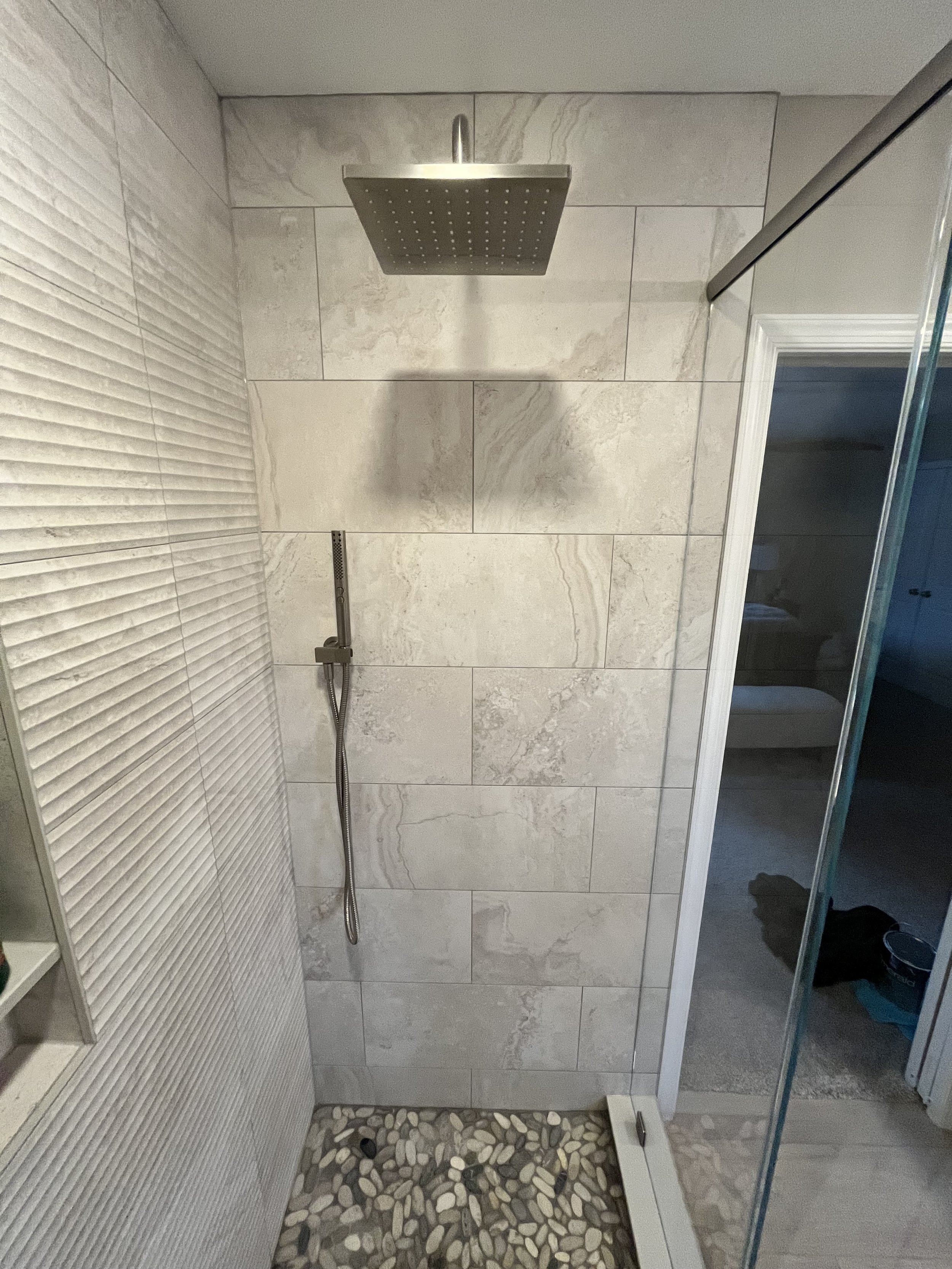A walk-in shower with a large rainfall showerhead, a handheld shower wand, beige marble tiles on the wall, and pebble stones on the shower floor.