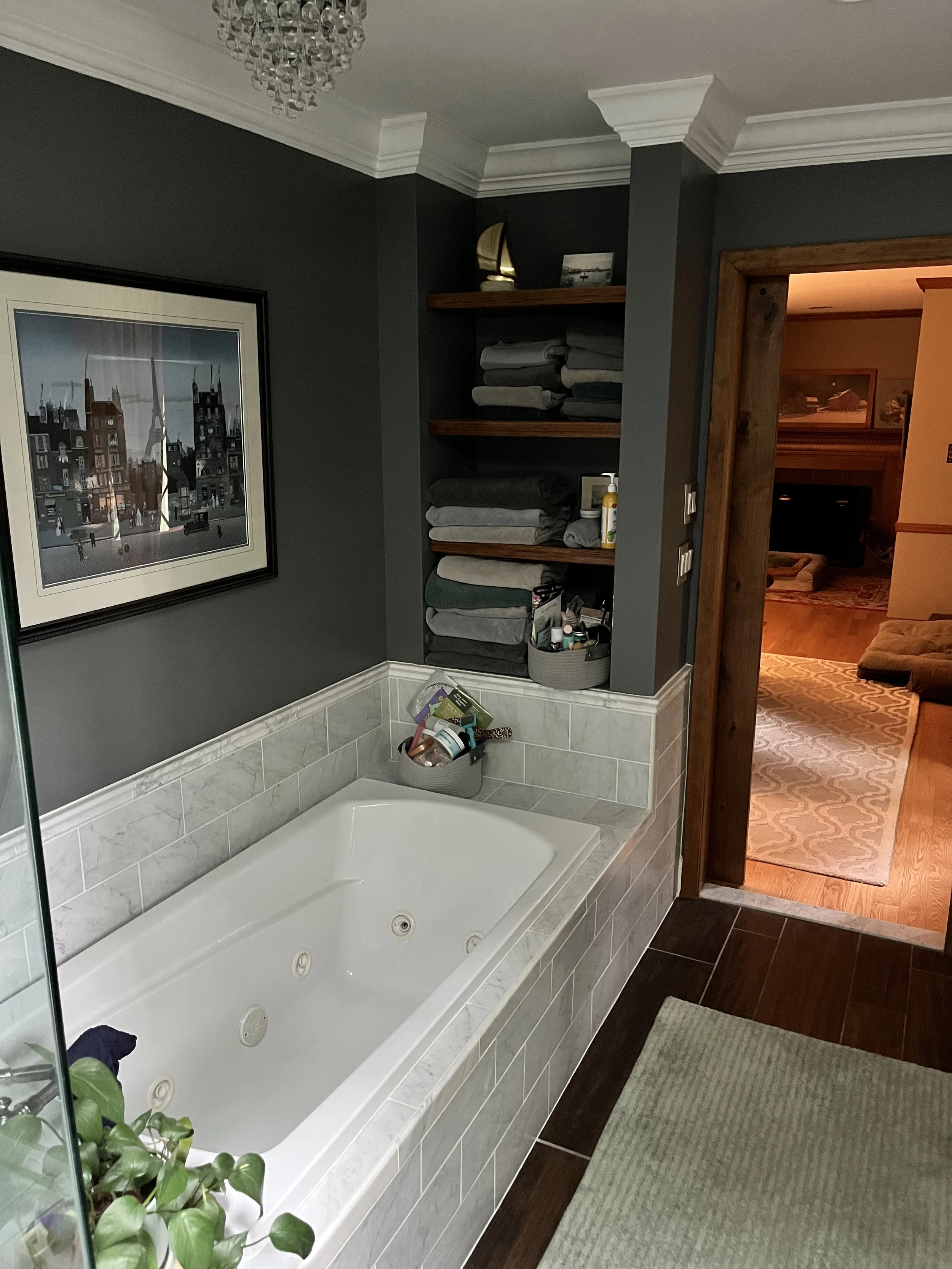 A bathroom with a built-in jacuzzi tub, dark gray walls, white crown molding, wooden shelves with folded towels, a framed picture of city buildings, and a partially visible living room with a beige rug and an orange sofa.
