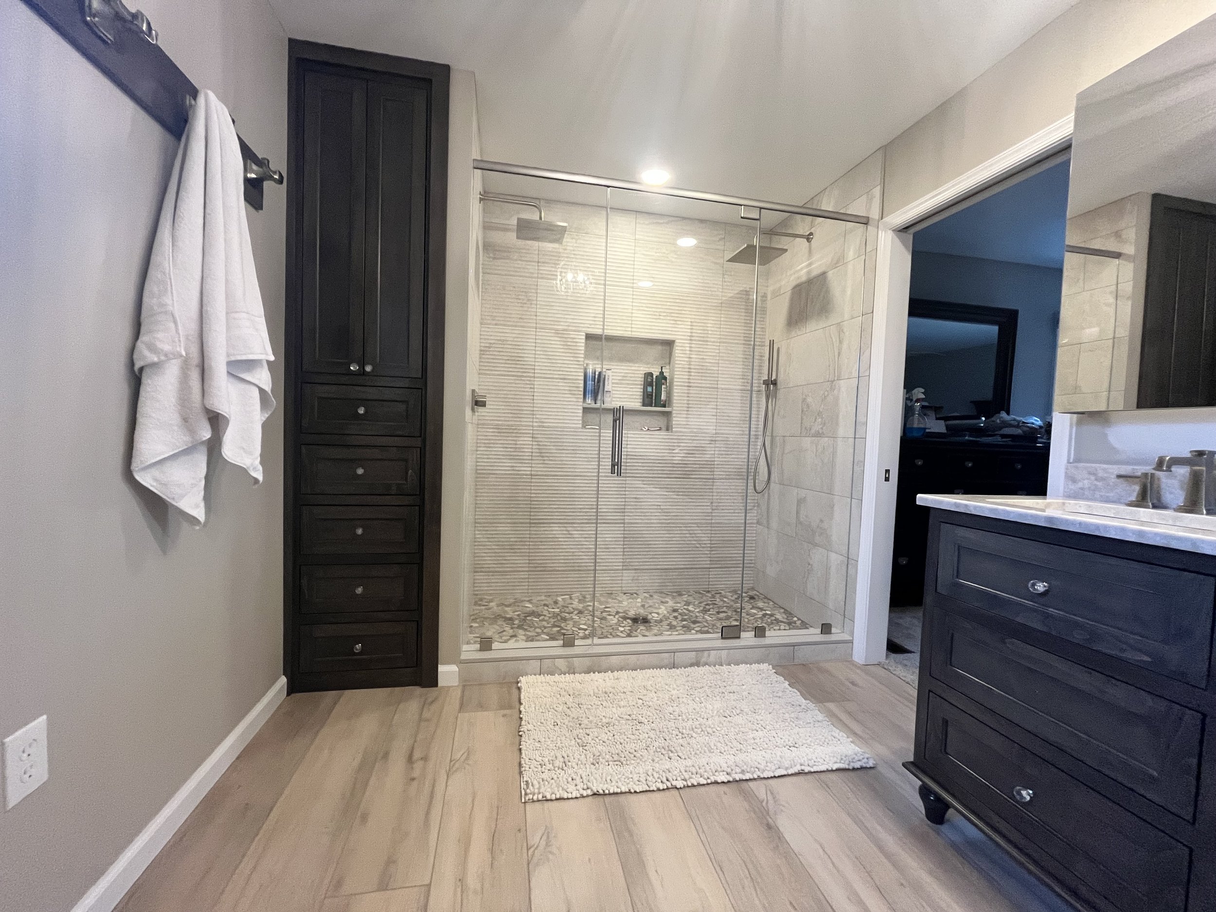 A bathroom with a glass-enclosed shower featuring a rainfall showerhead, dark wooden cabinets, a white fluffy rug, and a small counter with a sink, mirror, and toiletries, with wood flooring.