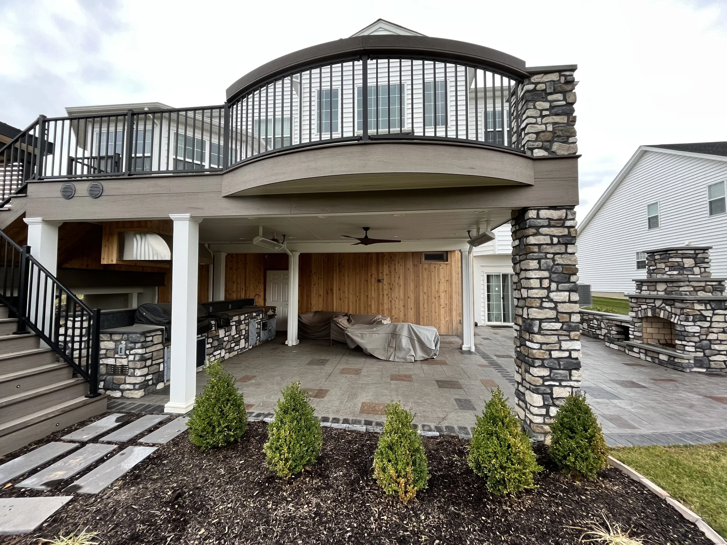 The image shows a backyard patio area under a house with a second-story balcony. The patio is paved with stone tiles and is decorated with four small bushes and a modern pathway leading to the patio. The house features a stone column, a ceiling fan, 