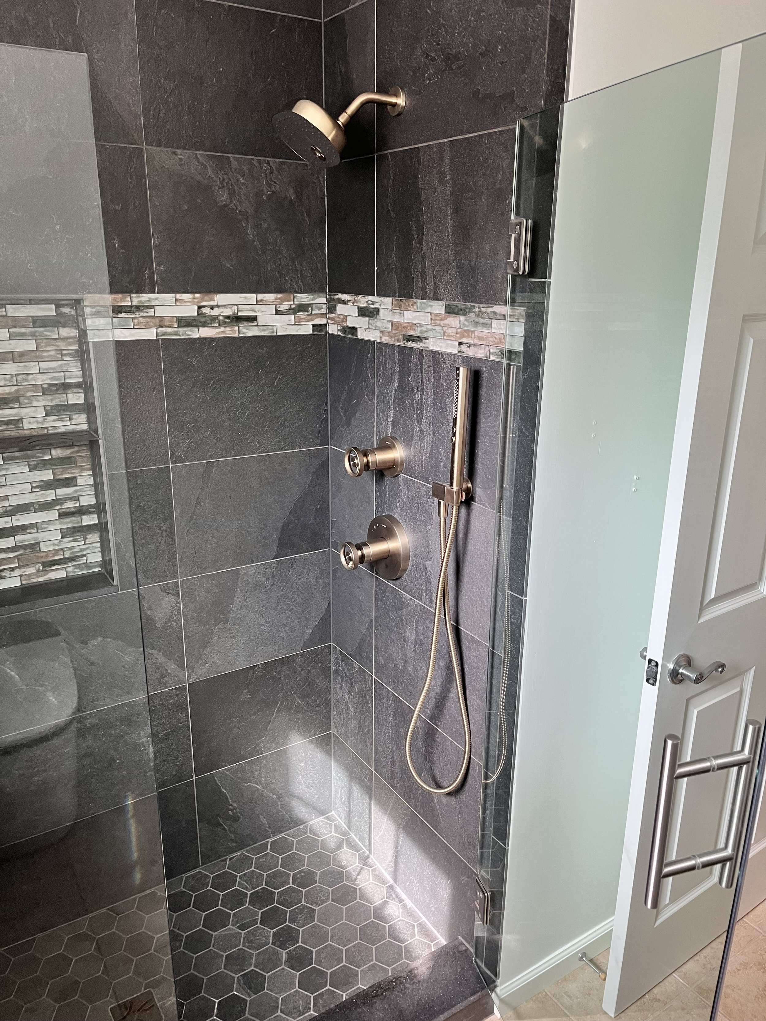 Shower with dark gray tiles, a decorative mosaic strip, a stainless steel shower head, a handheld shower wand, and a glass door, in a bathroom.