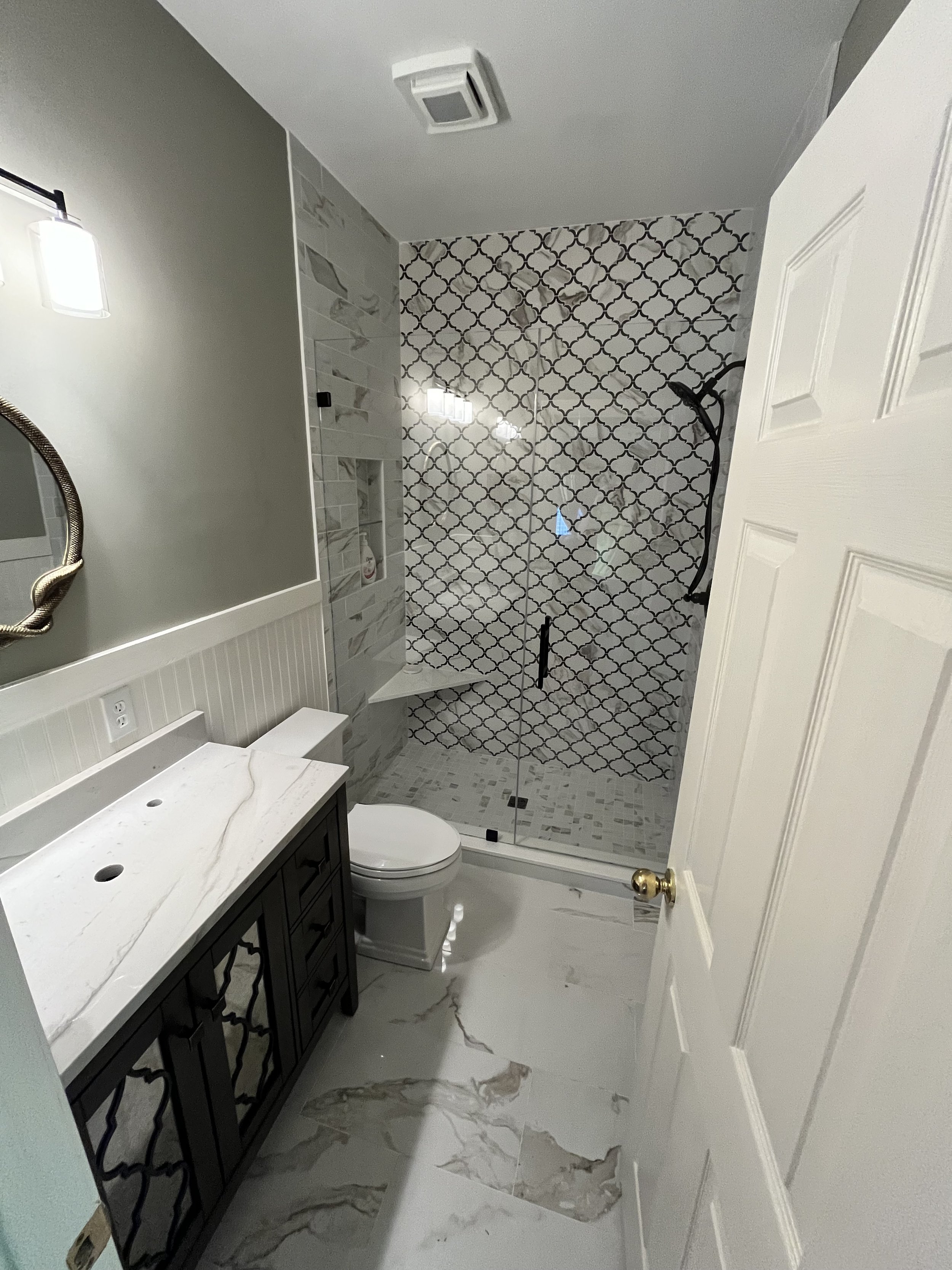 A modern bathroom with a marble floor, double-sink vanity with black cabinetry and marble countertop, mirror, and gray wall above beadboard wainscoting, and a glass-enclosed shower with patterned tiles and a rain shower head.