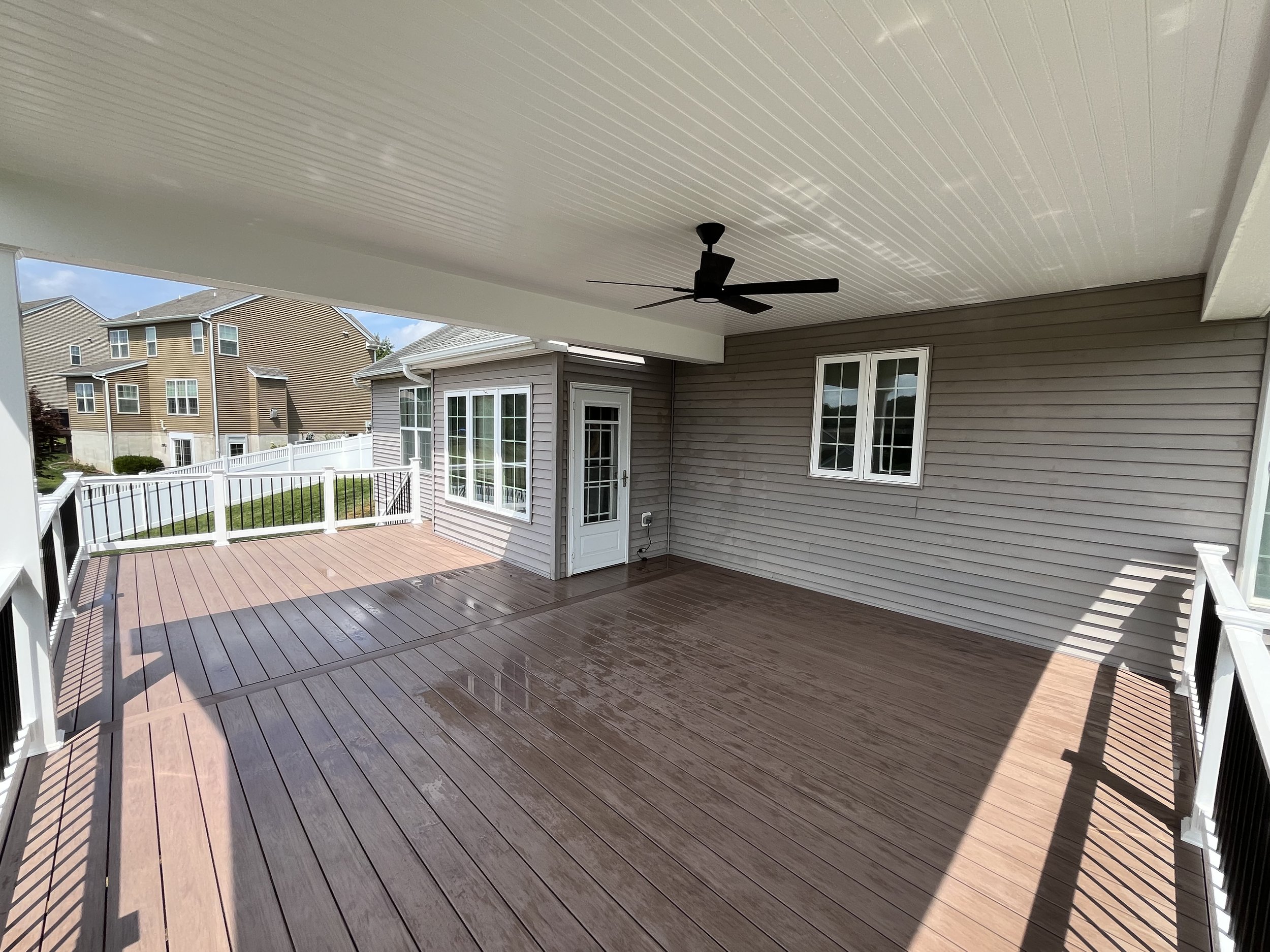 Covered backyard patio with wooden flooring, white railings, and a ceiling fan, adjacent to beige and gray houses.