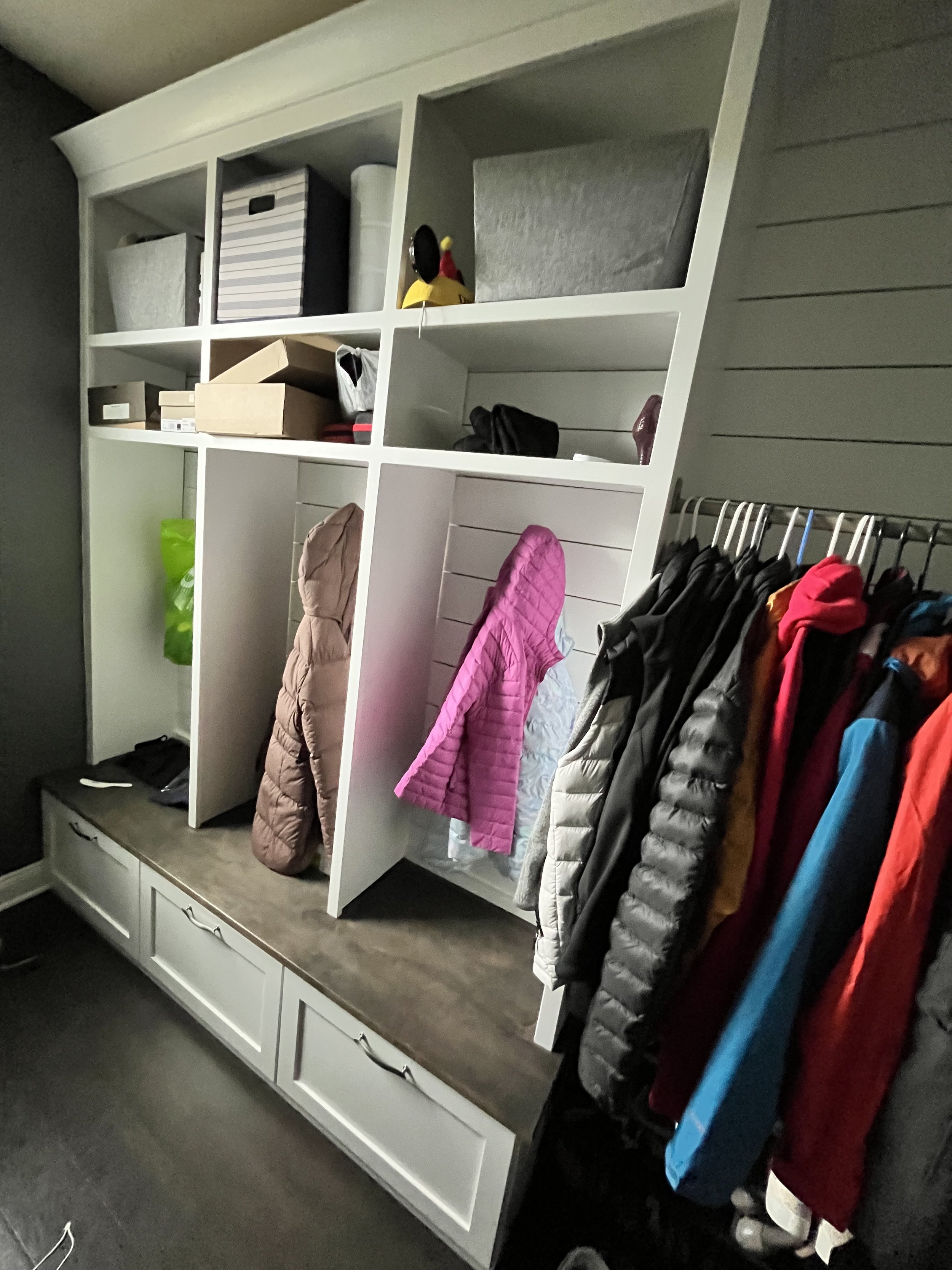White storage cubby with hooks holding jackets and coats in various colors, countertop with boxes and miscellaneous items, and gray wall in the background.