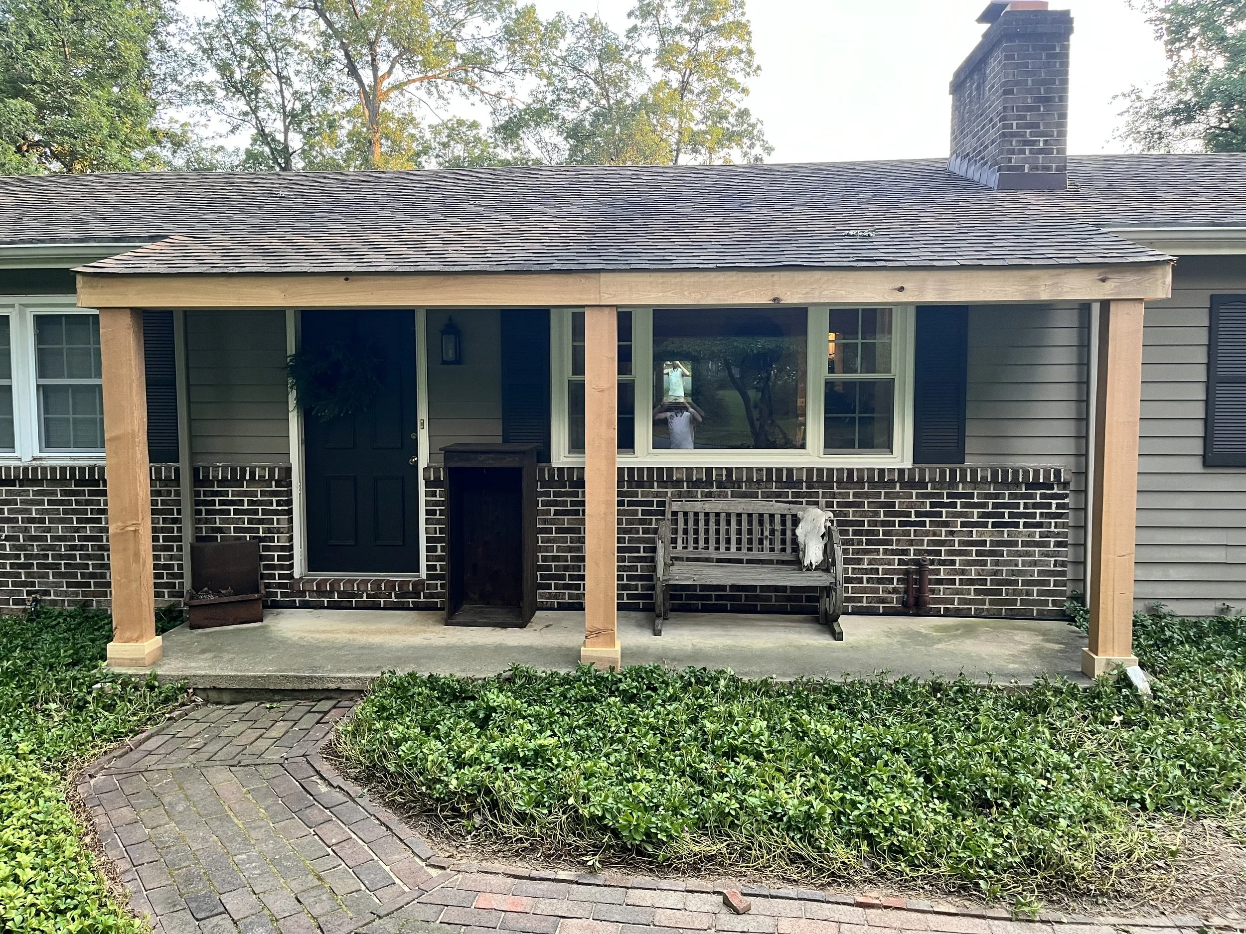 A house with a porch area under construction, with two wooden posts and a wooden beam, a sheltered area with a bench and decorative animal skull, large windows, a brick chimney, and greenery in the front yard with a brick pathway.