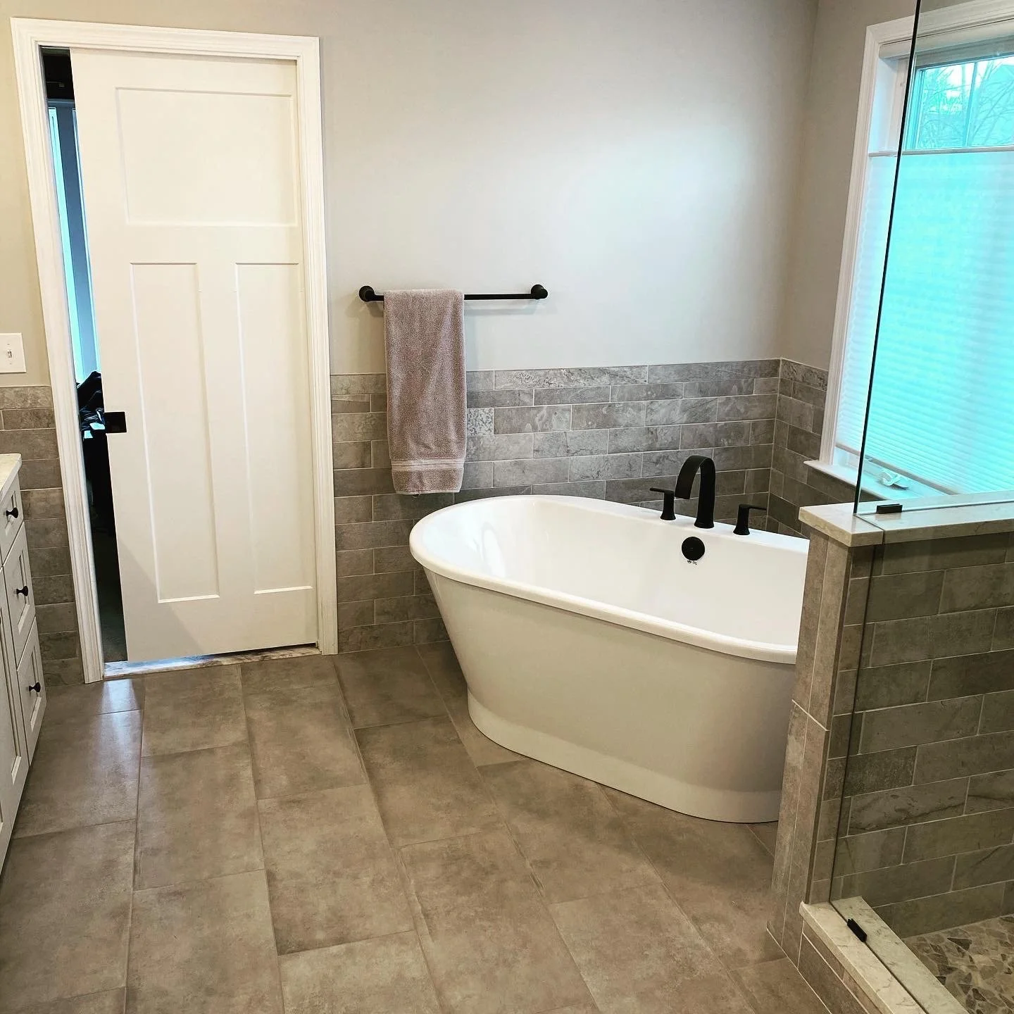 Bathroom with a white bathtub, grey tiled walls, a towel hanging on a black towel bar, a window with blinds, and a door.