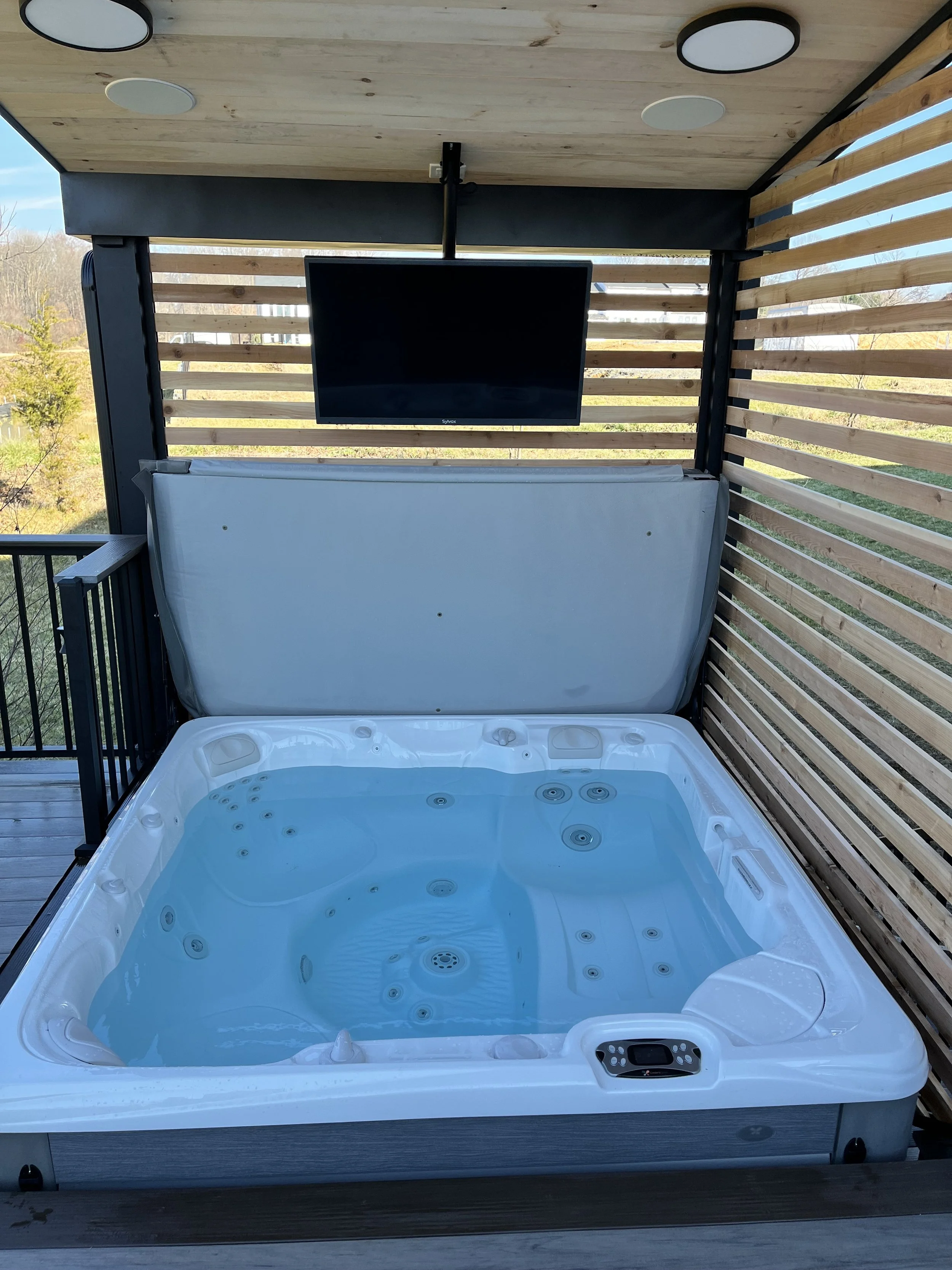 A hot tub filled with water on an outdoor balcony, with a mounted television and wooden slat privacy screen.