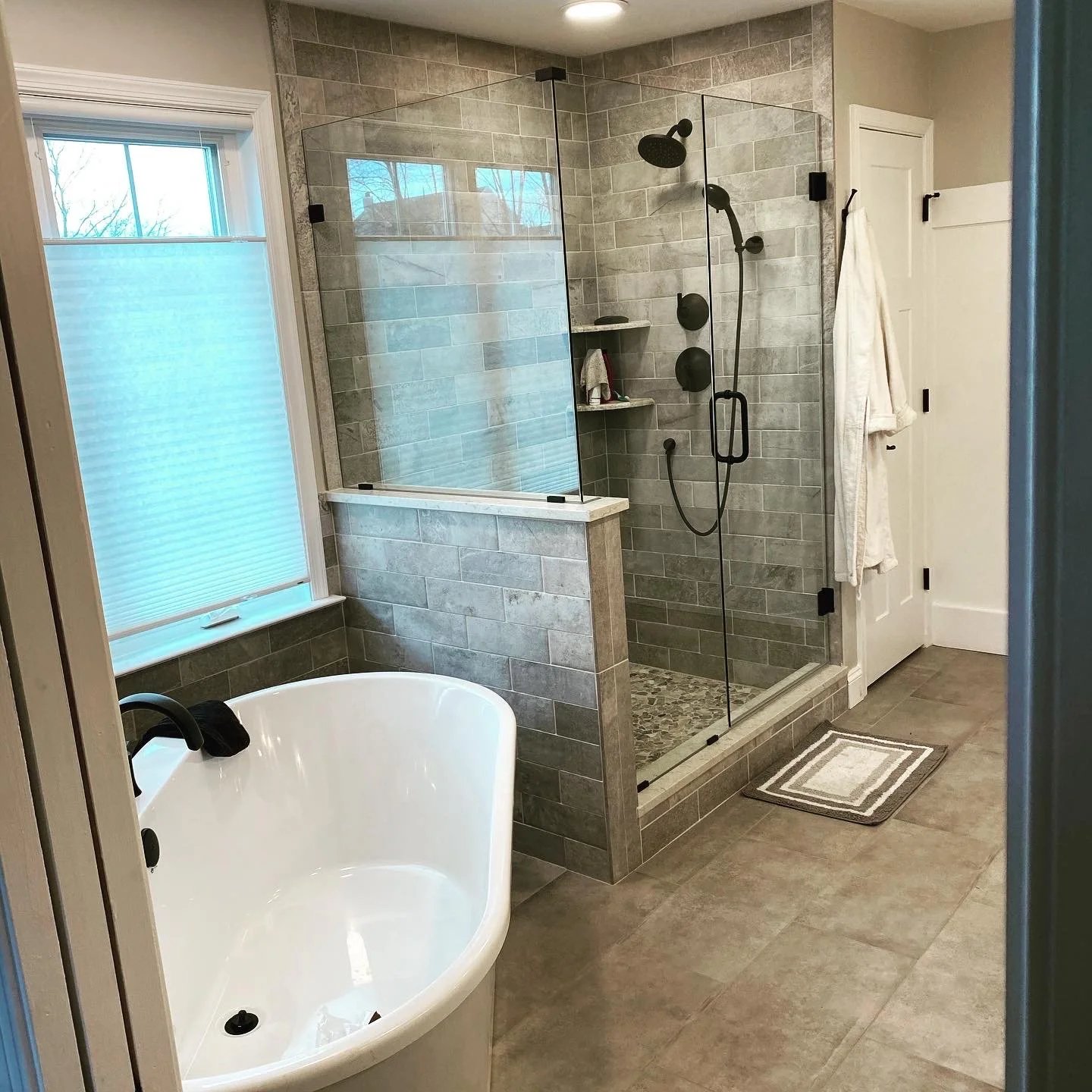 Modern bathroom with a freestanding bathtub, a glass-walled shower with black fixtures, a window with blinds, and neutral-colored tiles.