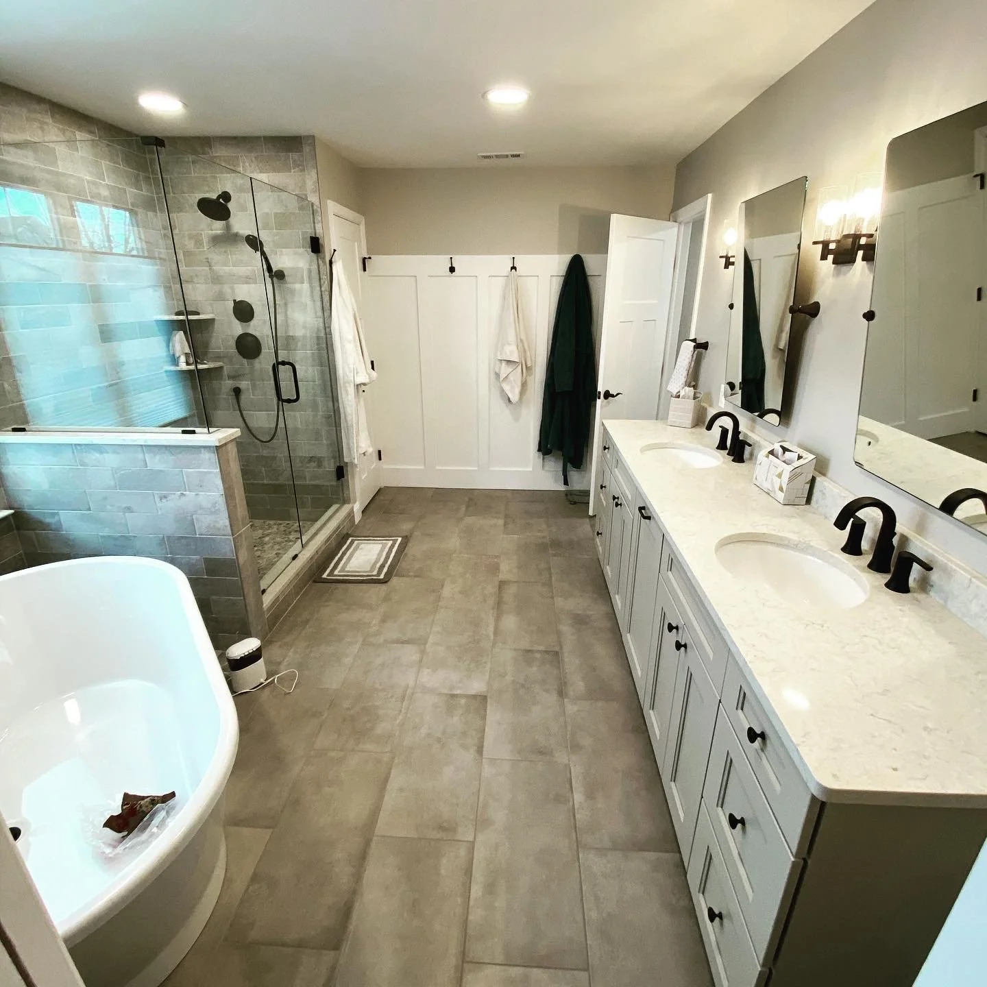 A modern bathroom with a bathtub, a glass-enclosed shower, a double vanity with two sinks, and large mirror panels. There are black fixtures and fixtures, beige tile flooring, and a light gray wall with decorative elements.