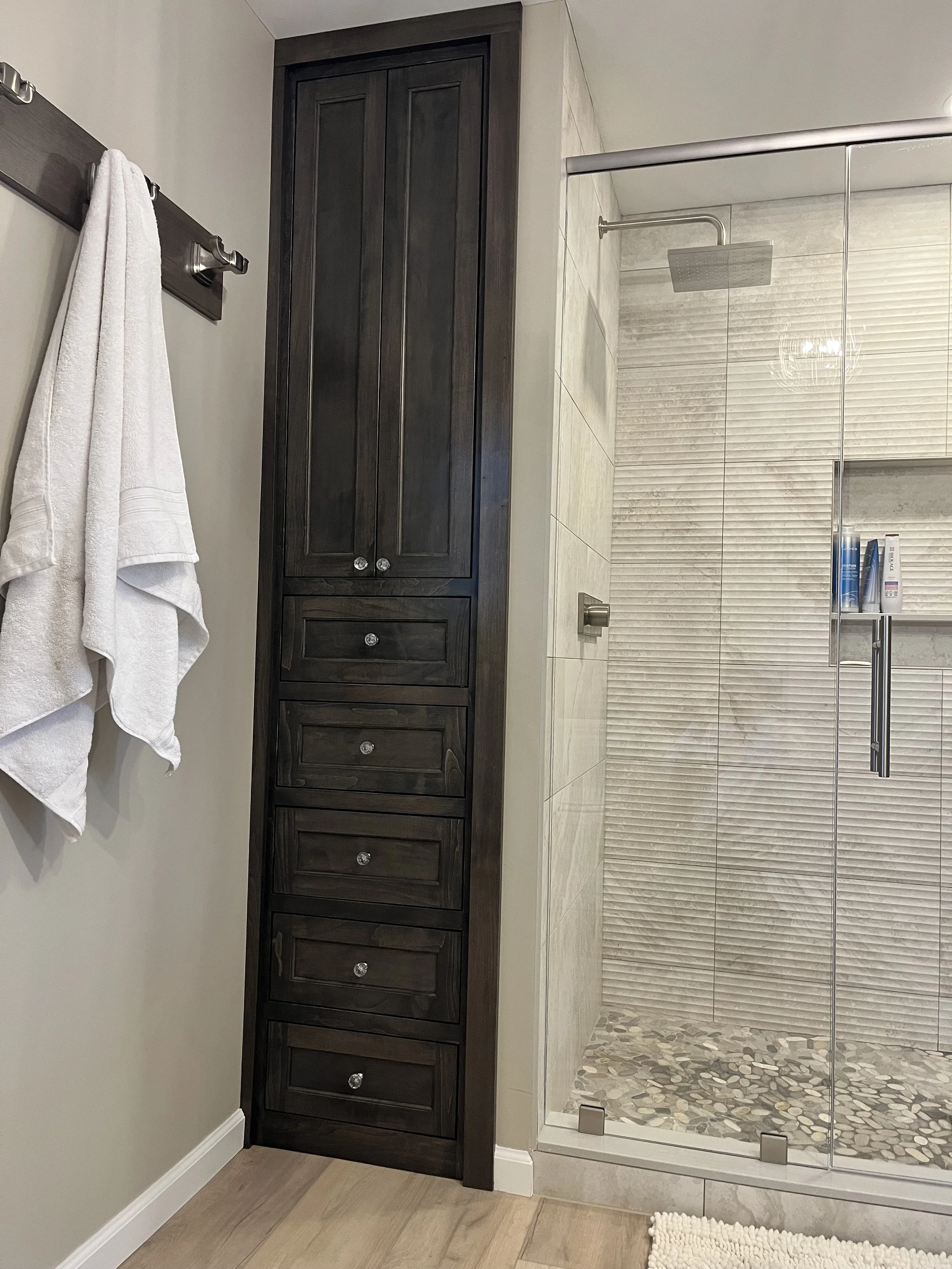 A bathroom with a dark wood bathroom cabinet against a beige wall, a glass-enclosed shower with pebble flooring, a shower head, and bottles of shampoo and body wash inside the shower.