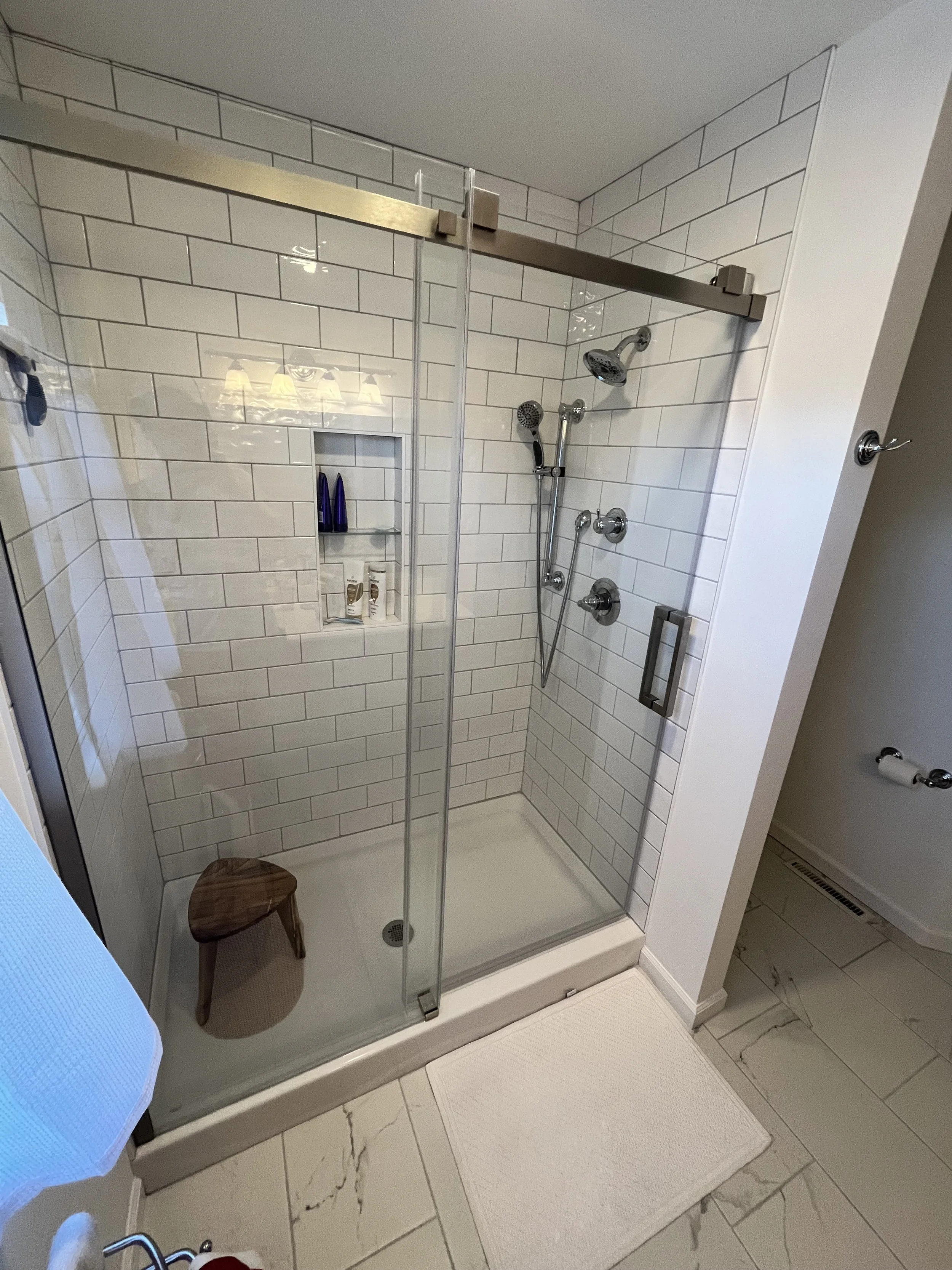 Shower with white subway tile walls, glass door, and built-in shelf holding toiletries. Small wooden stool inside shower. White bath mat outside on tile floor.
